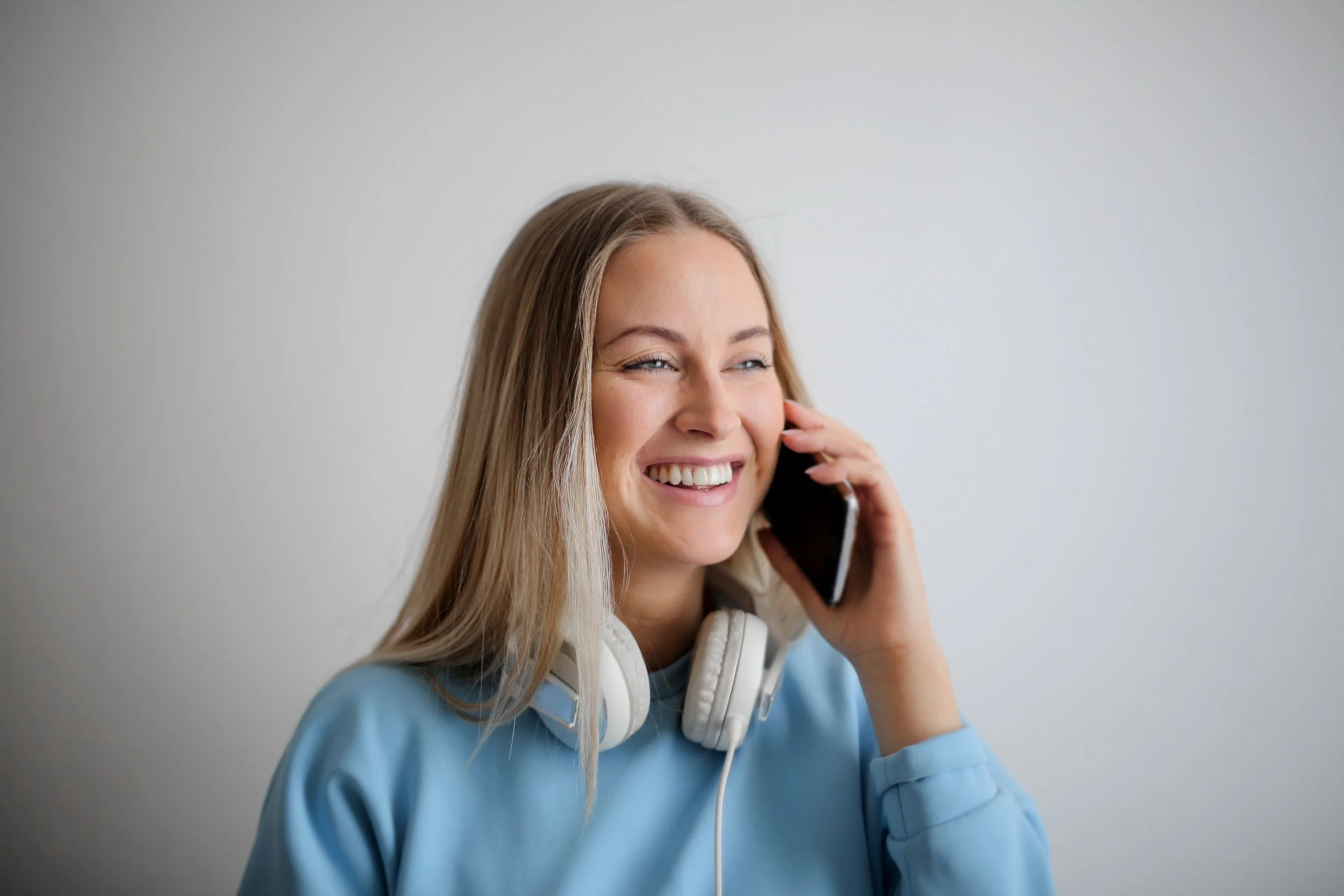 Young woman with long blonde hair smiling, talking on a cellphone, wearing headphones around her neck, and a light blue hoodie.