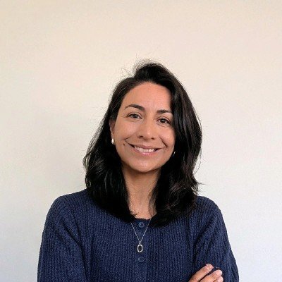 A woman with dark hair smiling and wearing a blue sweater against a plain light-colored wall.