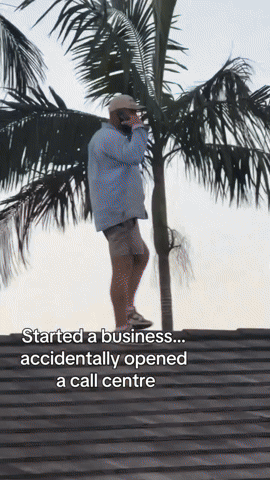 Person standing on rooftop with palm trees in background, talking on a cell phone, wearing a hat, sunglasses, shirt, and shorts. Caption: "Started a business... accidentally opened a call centre."