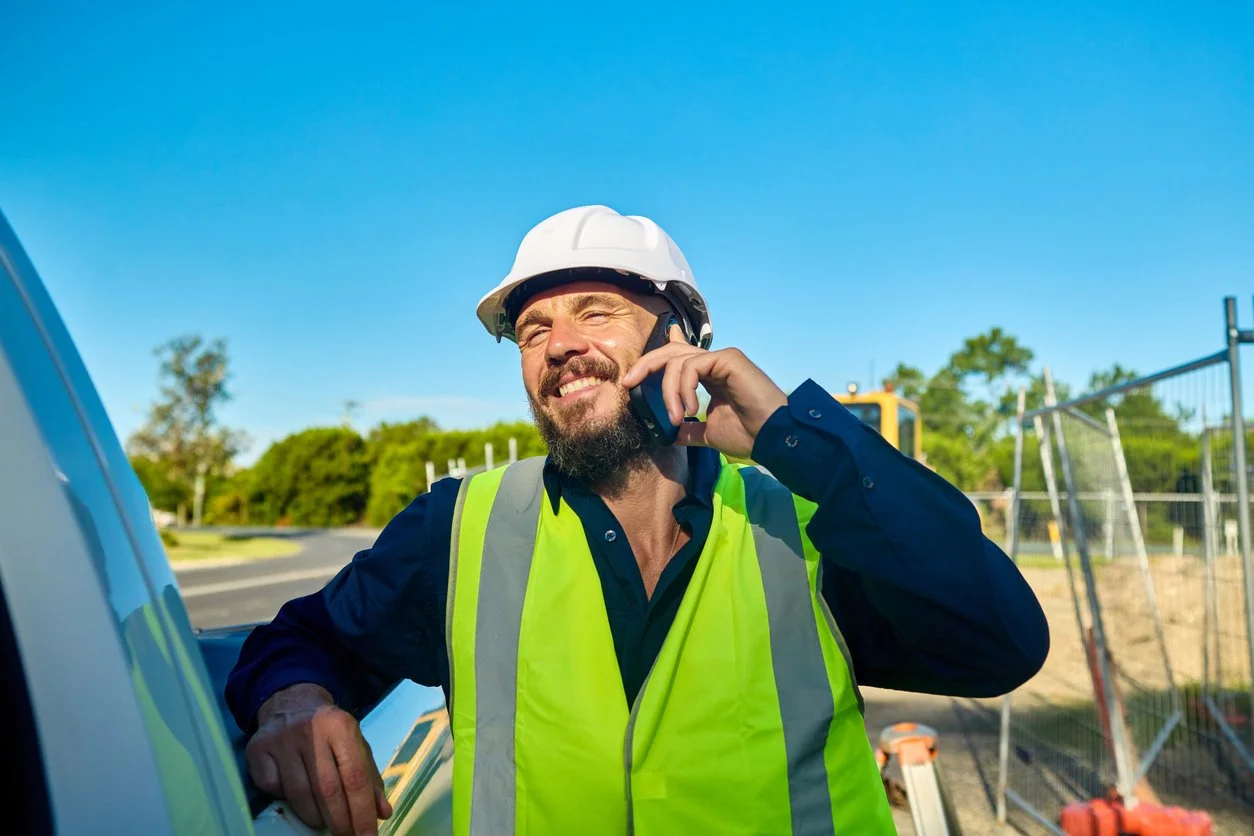A construction worker talking on a cell phone outdoors, wearing a white safety helmet and a yellow reflective vest, with trees and a fence in the background.