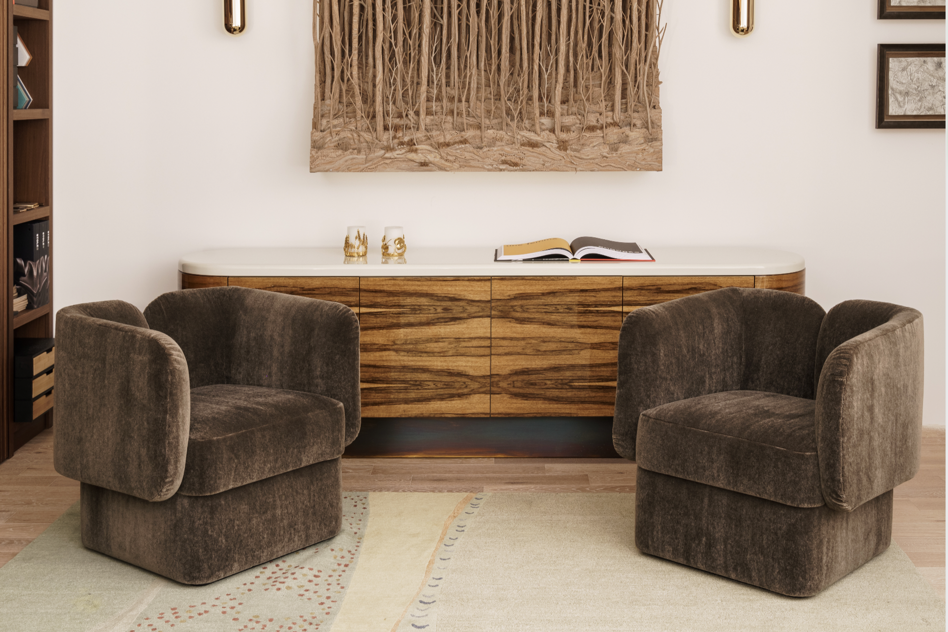 Living room with two brown velvet armchairs facing each other, a white and wood sideboard with decorative candles and a magazine on top, and a portion of a colorful area rug with geometric patterns.