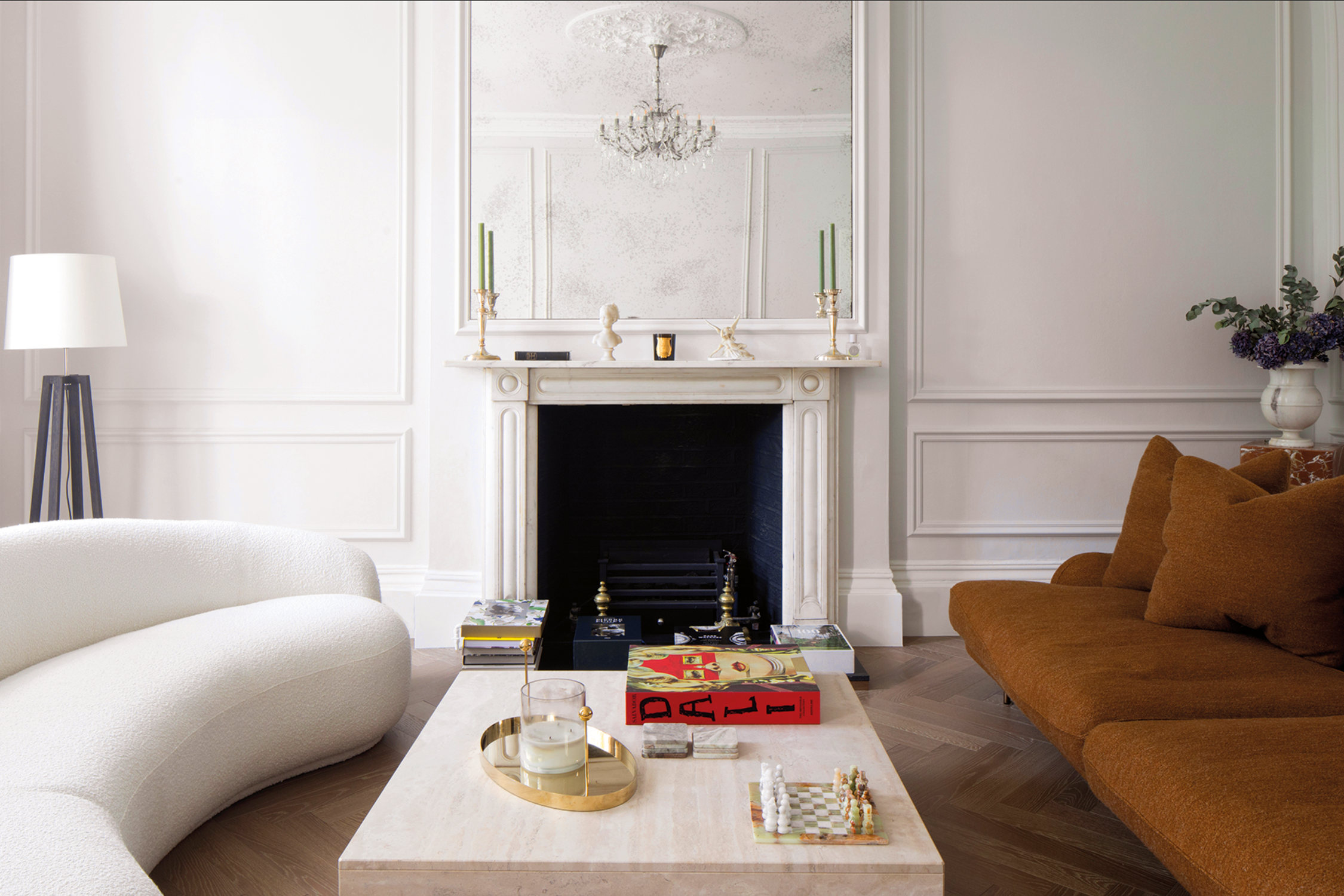 Living room with a fireplace, mirror, candles, and decorative items on the mantel. A white couch and a brown sofa are visible, as well as a white coffee table with books and a glass of water. A tall floor lamp is on the left, and a vase with purple f
