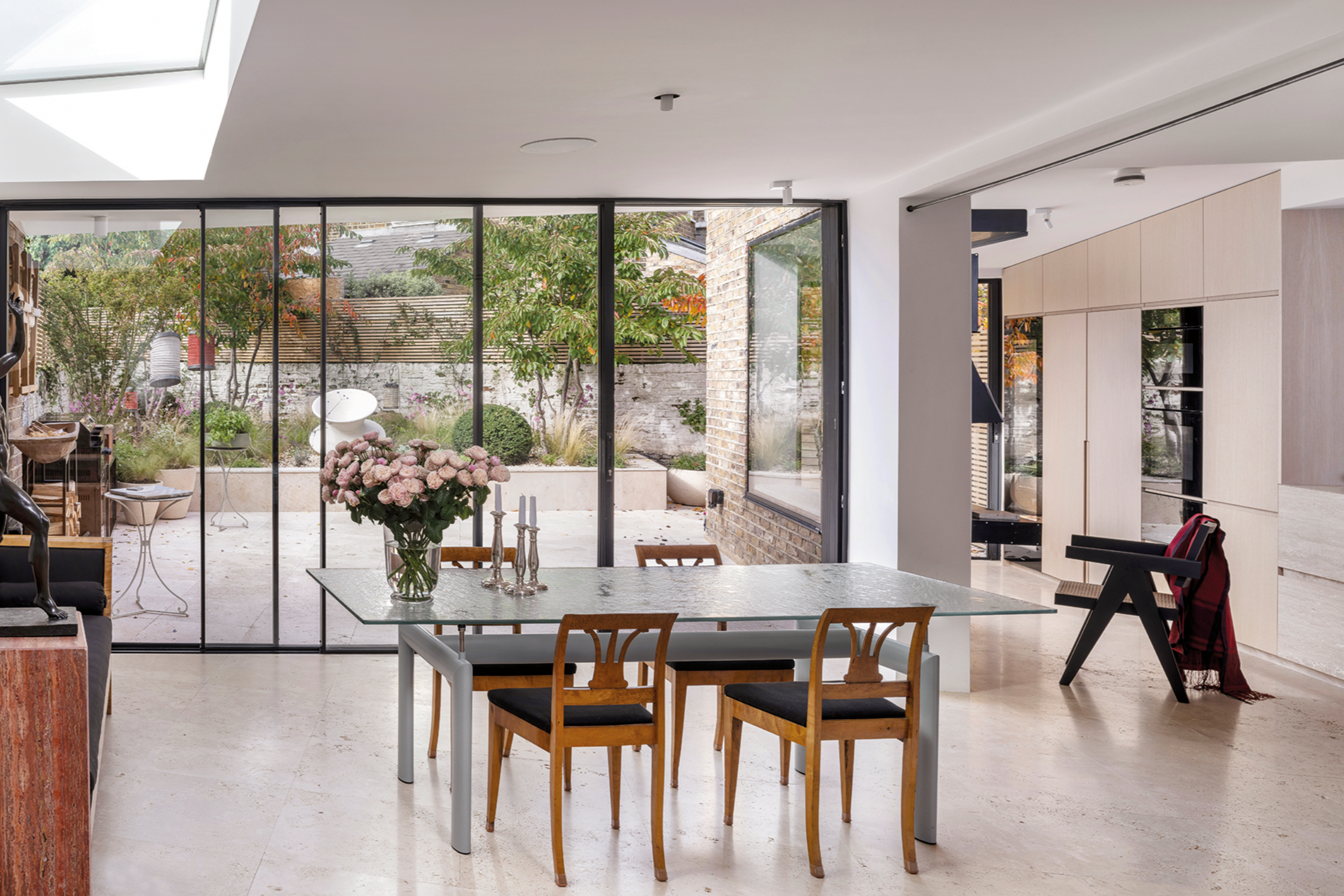 Modern interior dining room with a glass door opening to a garden. A white table with wooden chairs, pink flowers in a glass vase, and candles are visible.