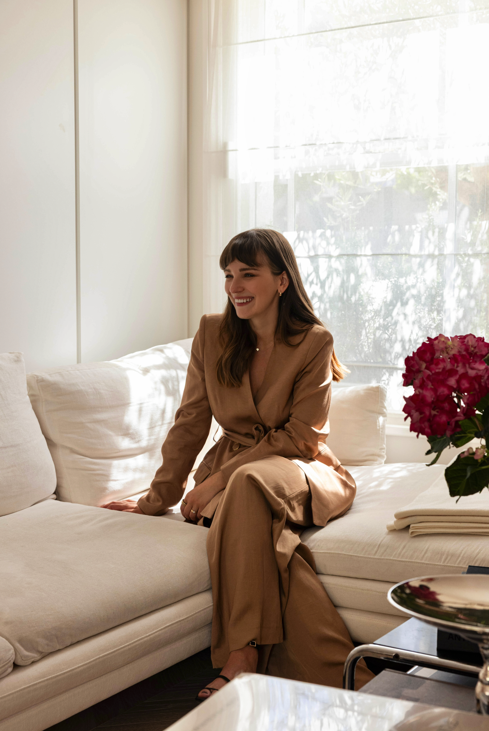 A woman with long brown hair and fair skin, smiling, sitting on a white couch in a bright room with large windows and sheer curtains, wearing a tan suit. There is a bouquet of pink hydrangeas on a table nearby.