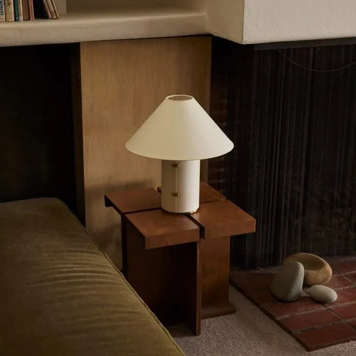 A white table lamp with a wide, conical lampshade sitting on a wooden nightstand beside a bed in a cozy room.