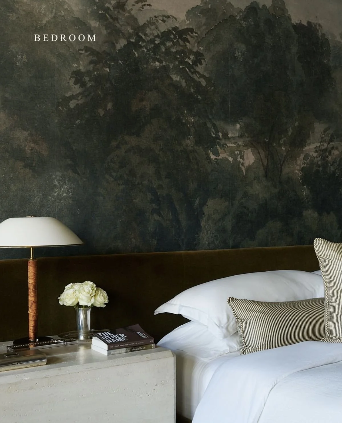 A bedroom with a dark mural featuring trees on the wall behind a bed. There is a white bedside table with a vase of white flowers, a few books, and a lamp with a white shade. The bed has white linens and pillows, with some striped pillows.