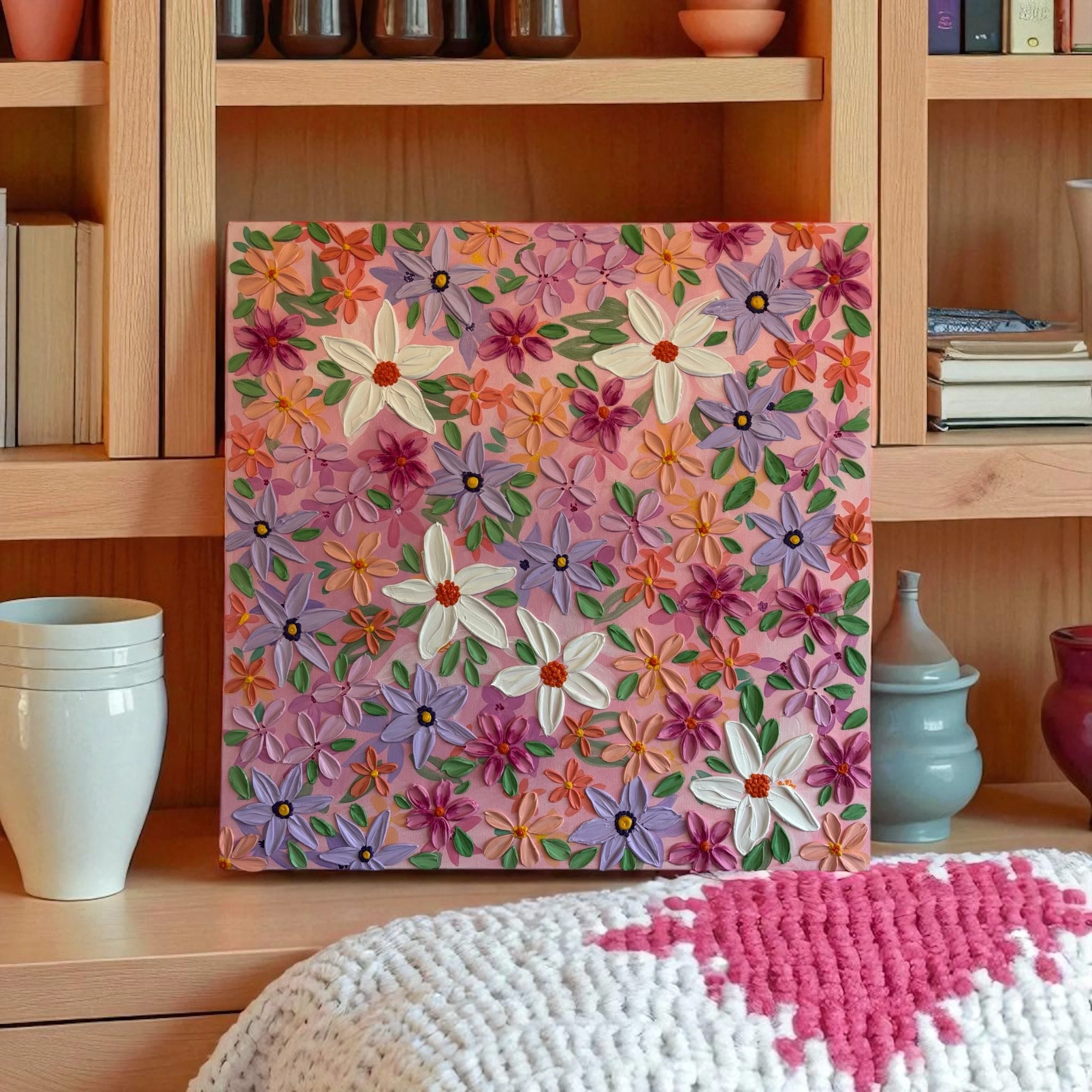 A colorful painting of various flowers in shades of pink, purple, white, and orange on a pink background, hung on a wooden bookshelf.