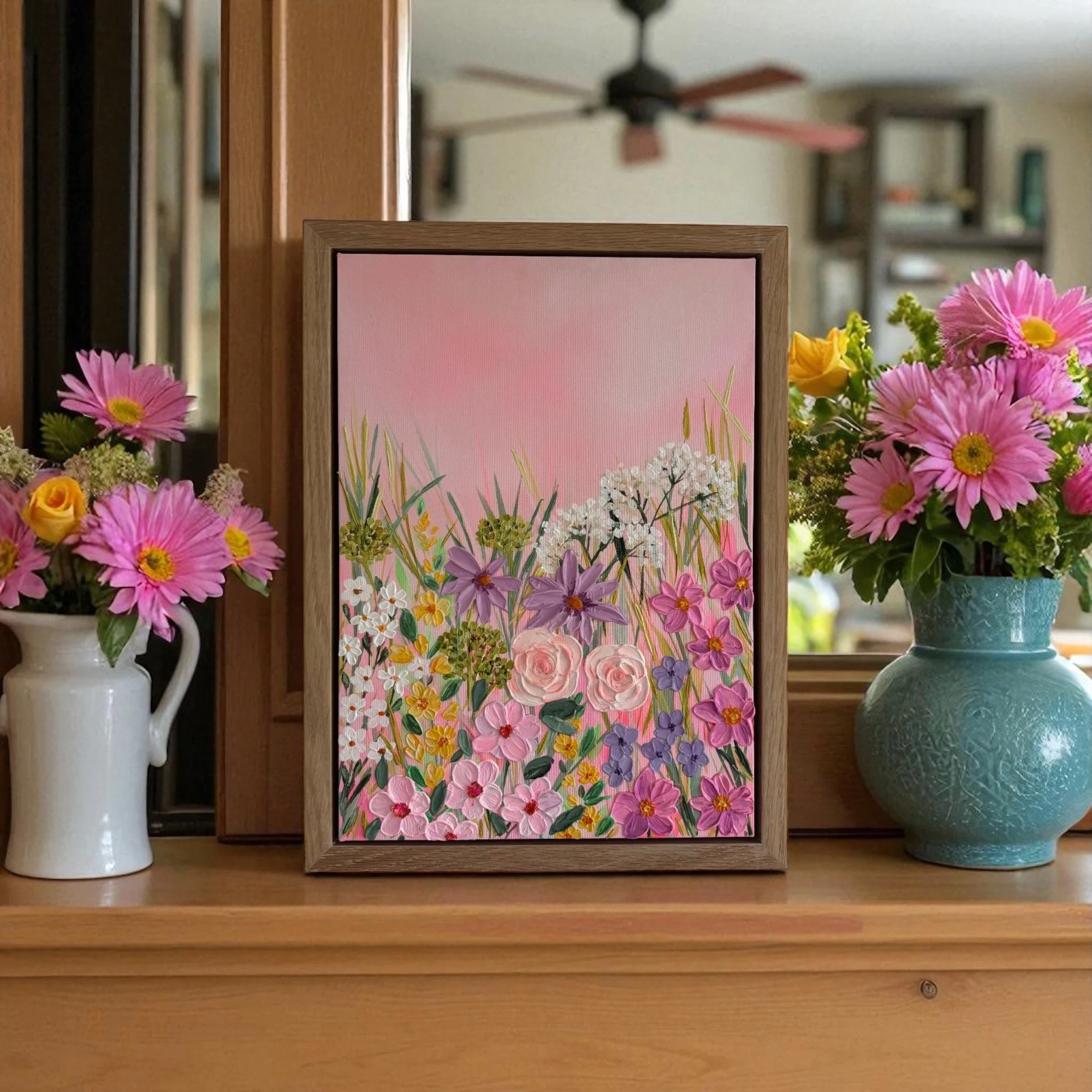 A framed floral artwork surrounded by pink and yellow flowers, placed on a wooden shelf with a white pitcher on the left and a gray vase on the right.