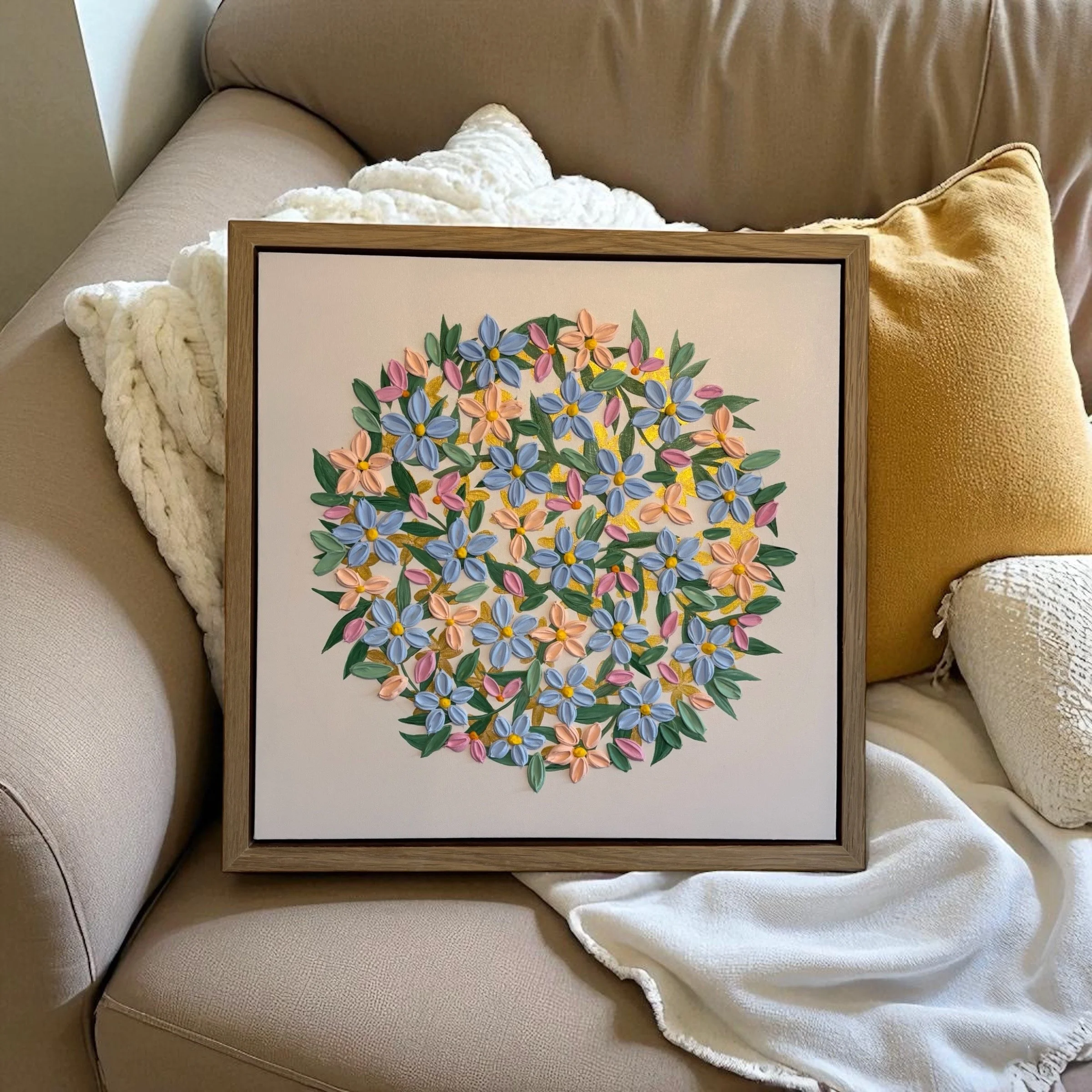 Framed floral painting of pink, purple, yellow, and blue flowers on a beige couch with a white and yellow blanket and a beige pillow.