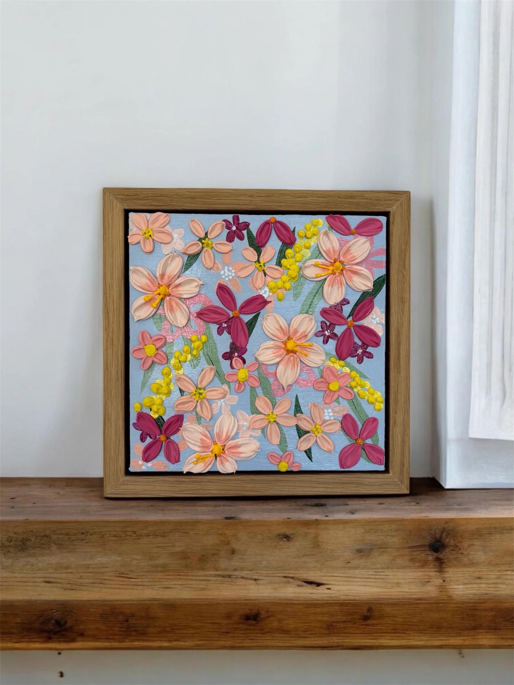 A framed floral artwork featuring pink, purple, and yellow embroidered flowers against a light blue background, placed on a wooden surface near a white wall and curtain.