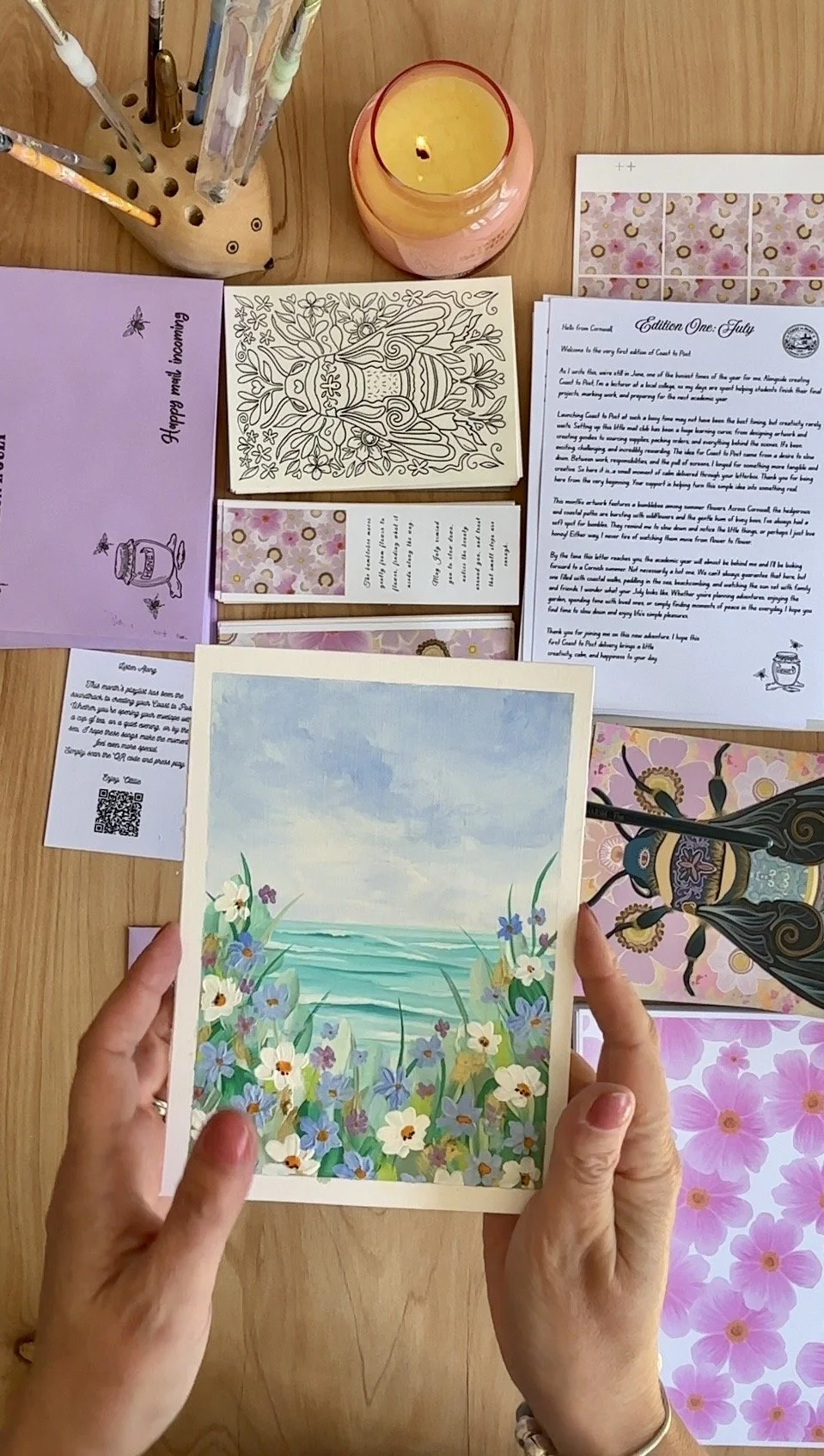 Hand holding a colorful illustrated card featuring a beach scene with flowers, ocean, and sky. The table in the background has various items including a candle, coloring pages, sheets with writing, and decorative papers, creating an artistic or craft workspace.