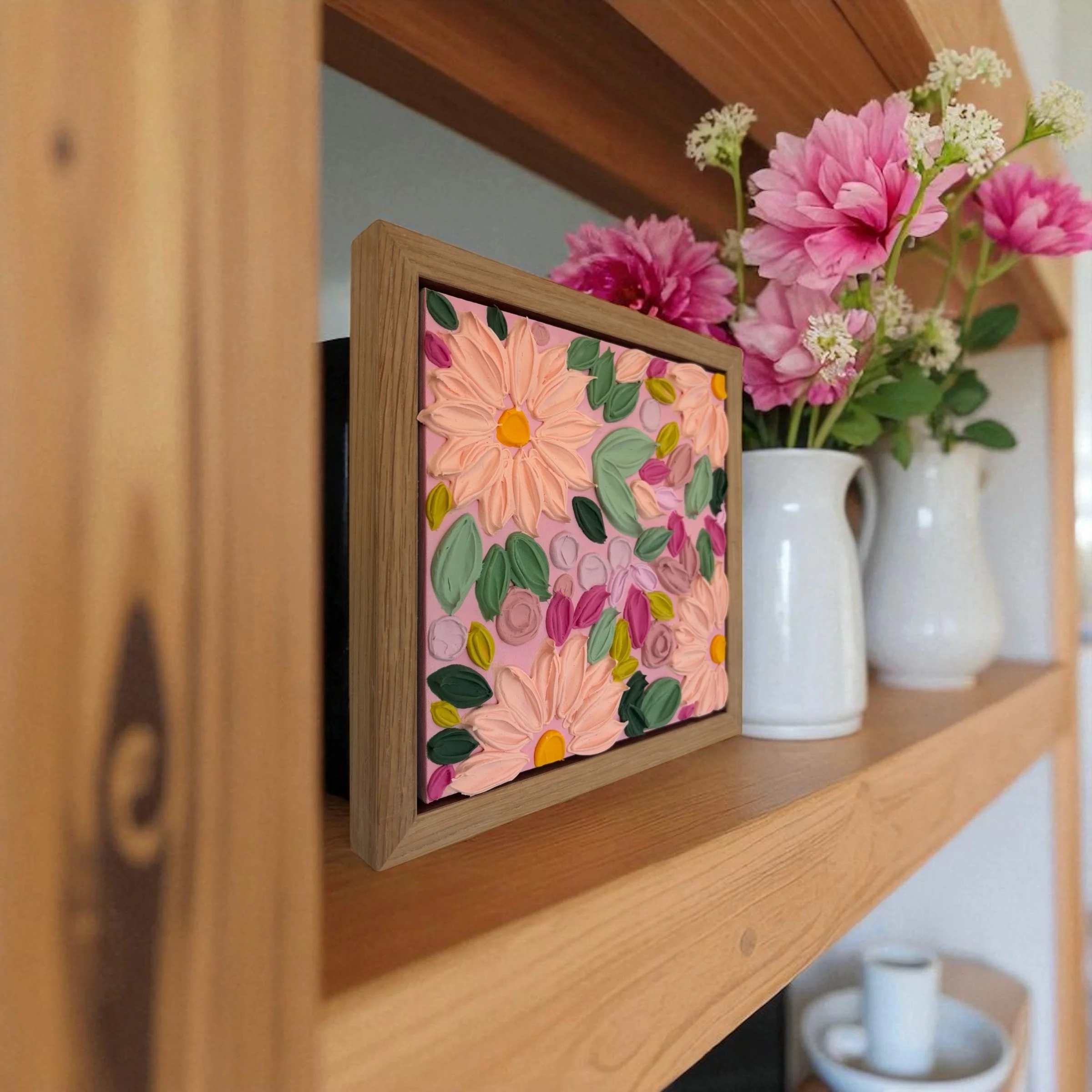 A wooden picture frame with a colorful floral painting, placed on a wooden surface next to a ceramic vase filled with pink and white flowers.