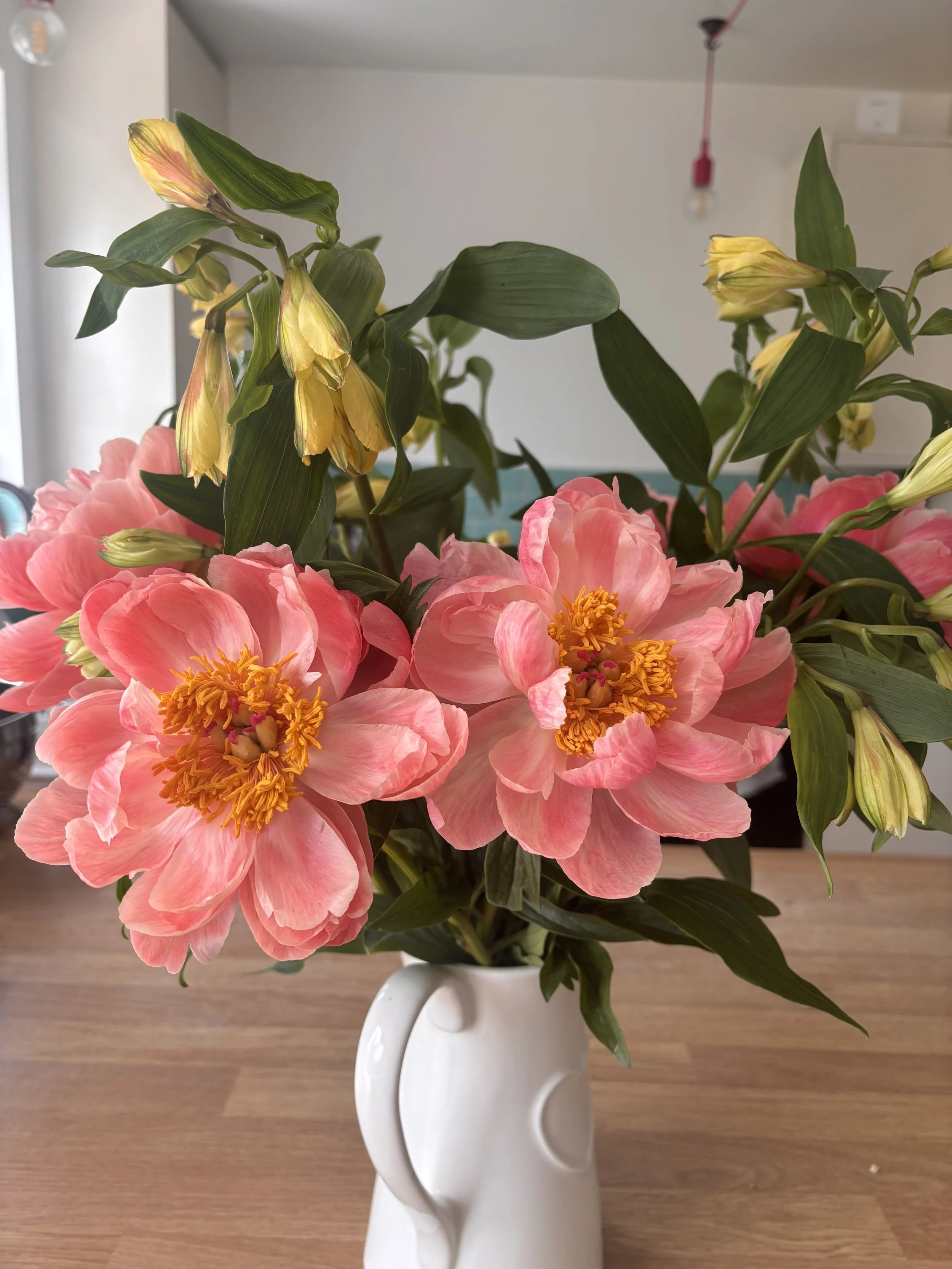A white ceramic vase with a unique curved handle design holds a bouquet of pink and yellow flowers with green leaves, placed on a wooden surface indoors.