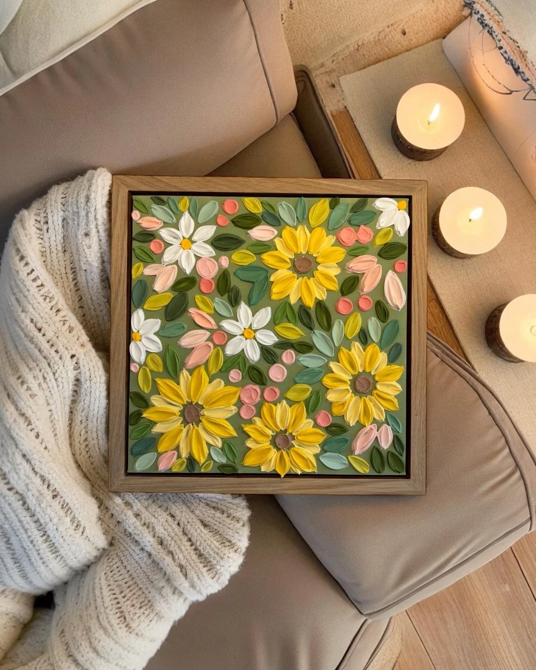 Framed floral mosaic art with yellow sunflowers, white daisies, pink roses, and green leaves, set on a beige couch next to three lit candles.