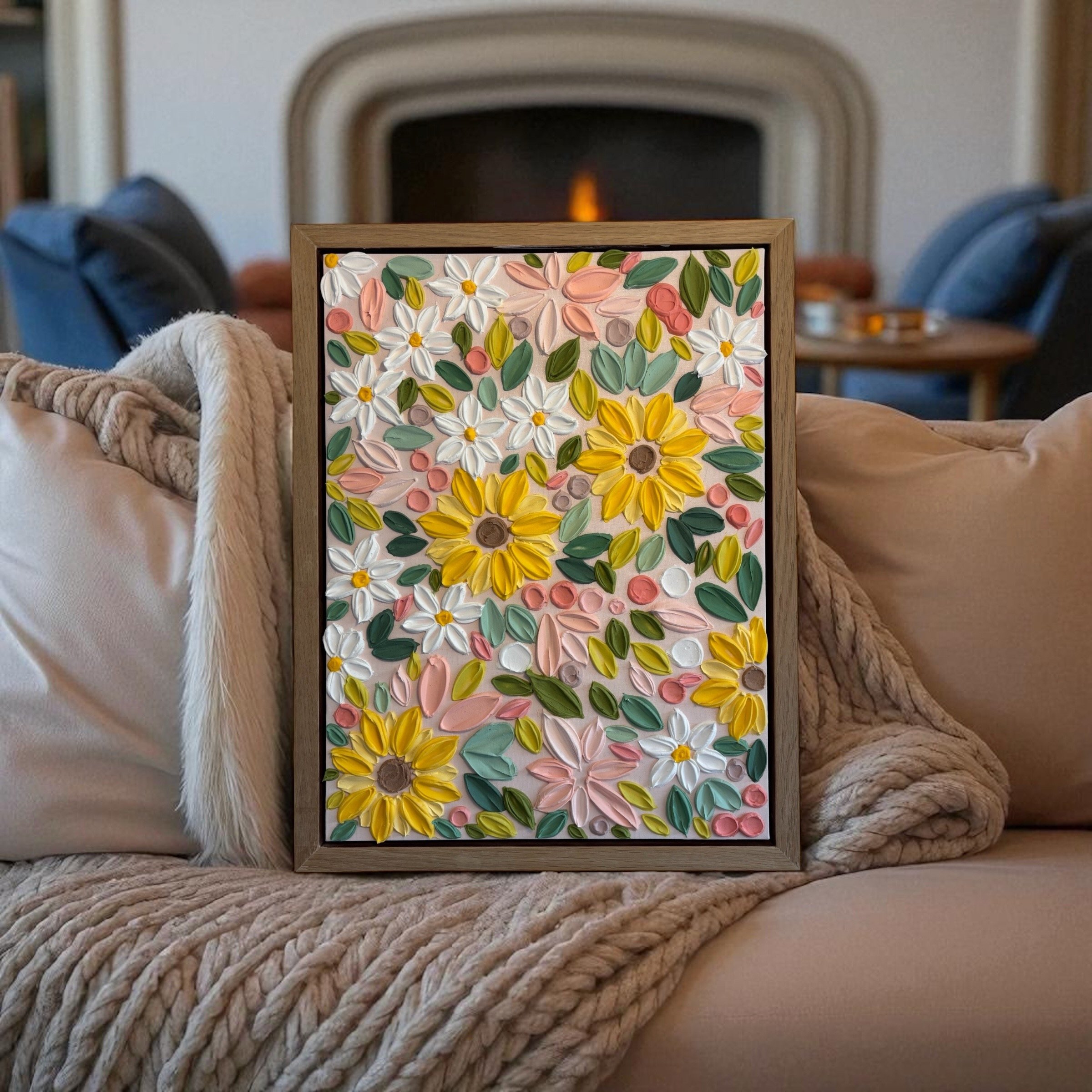 A framed cotton plant artwork with white, pink, yellow, and green textured petals and leaves, resting on a beige woven blanket in front of a fireplace.