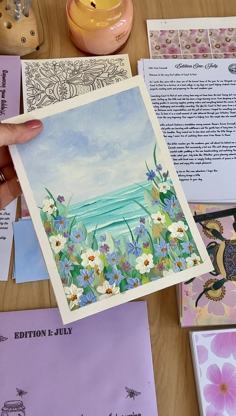 A hand holding a colorful illustration of a beach scene with blue water, sky, and flowers along the shore, surrounded by various papers, a candle, and other printed materials on a wooden table.