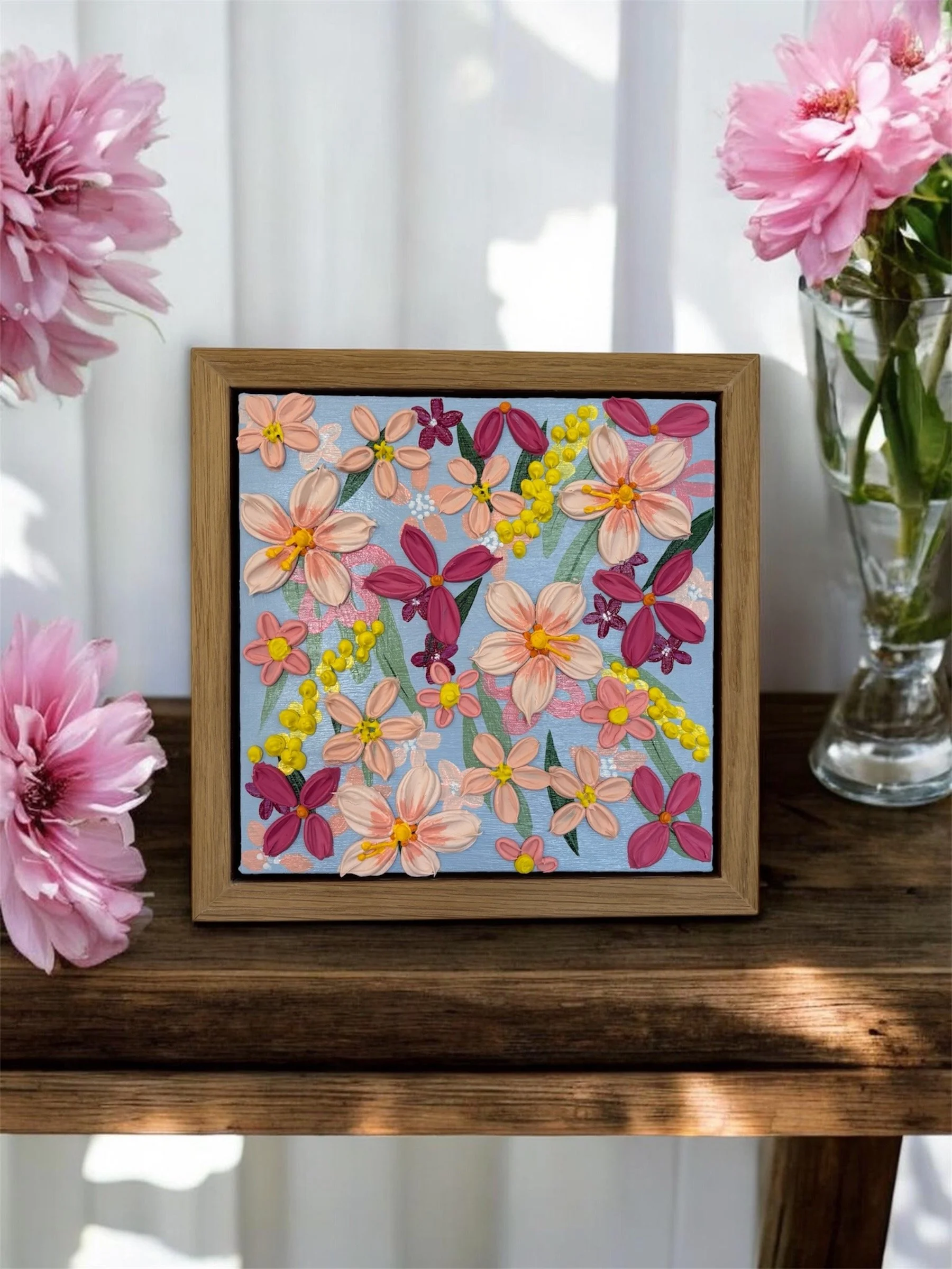 A framed embroidery artwork of colorful flowers on a blue background, placed on a wooden table with pink flowers in glass vases on either side.