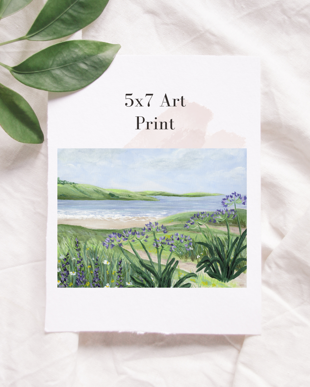 A printed art print titled "5x7 Art Print" featuring a landscape painting of a coastal scene with a beach, ocean, green hills, and purple flowers, placed on a white fabric with green leaves nearby.