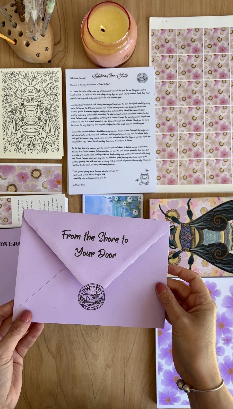 A person's hands holding a pink envelope with the text "From the Shore to Your Door" and a postal stamp. On the table are various postcards, a candle, a coloring page with a bee, and sheets of pink floral stickers.