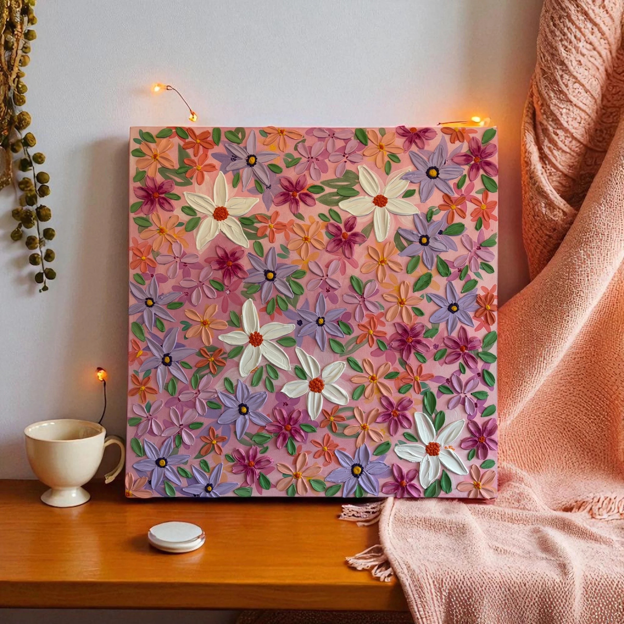 A handmade floral artwork with 3D painted flowers on a pink canvas, placed on a wooden surface. To the left of the artwork there is a string of small yellowish buds hanging down, and underneath is a white mug. To the right, a pink textured fabric is draped.