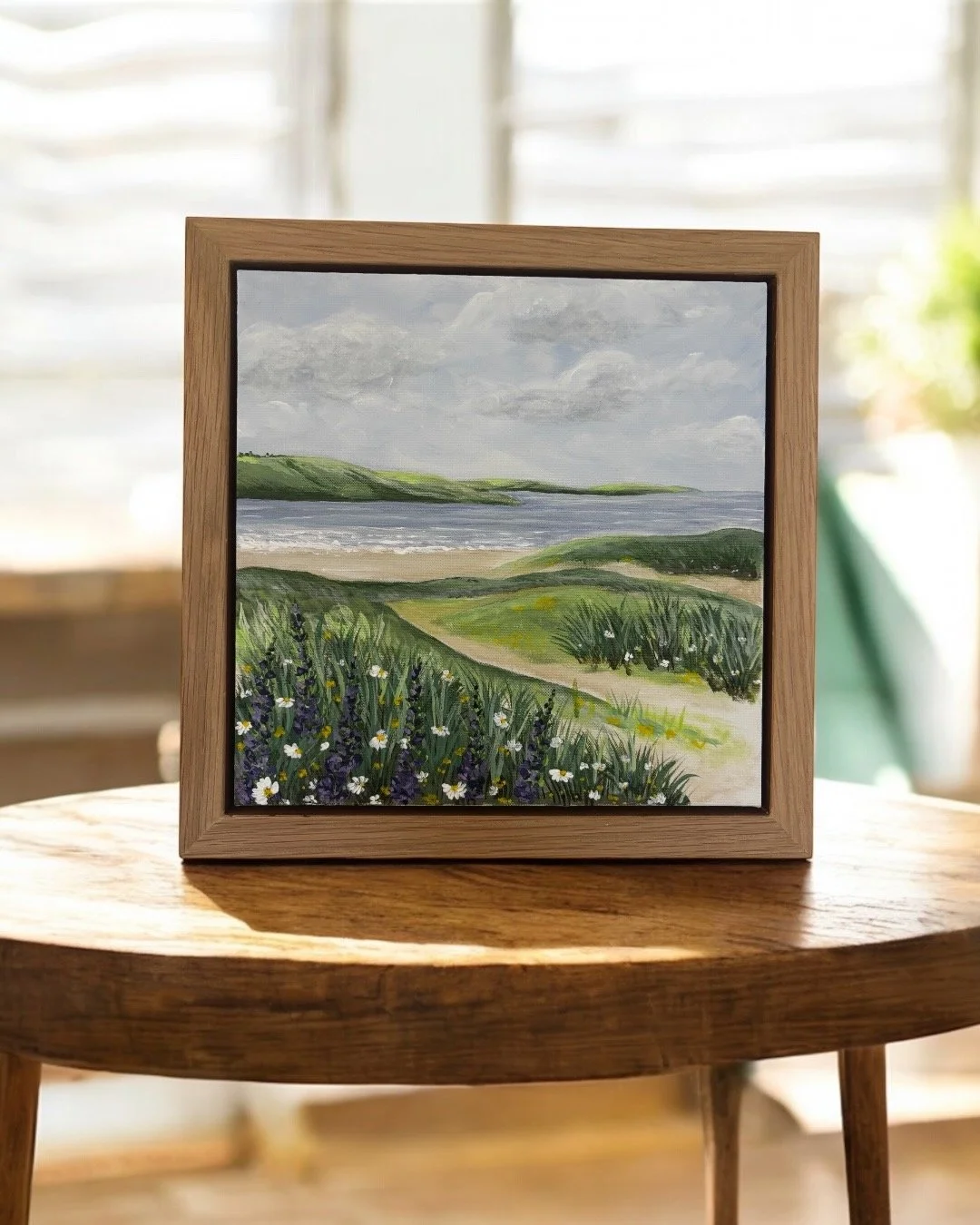 A framed landscape painting of green hills, a beach, and a cloudy sky, placed on a wooden table in a bright room.