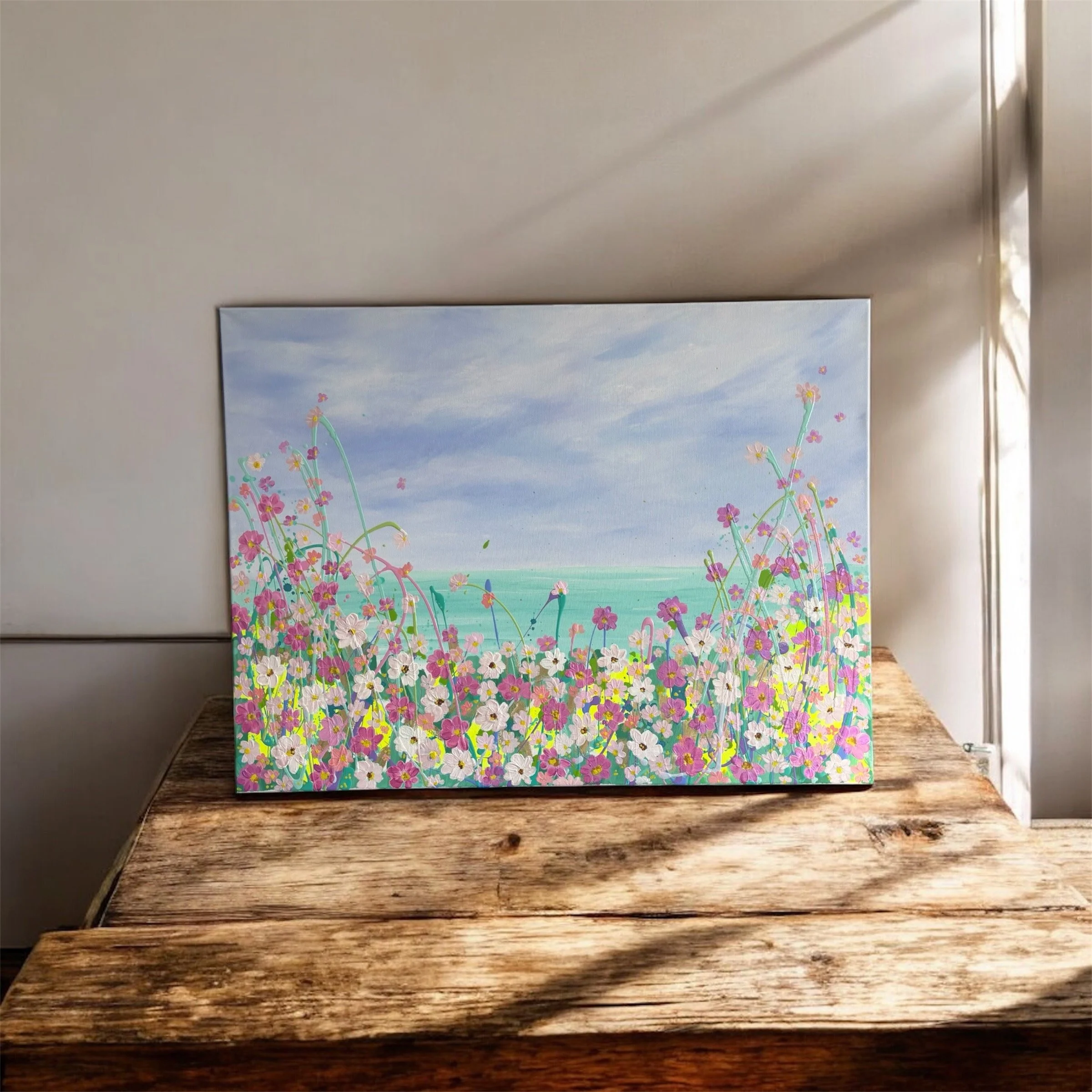 Colorful painting of a landscape with flowers on a canvas propped against a wall, placed on a rustic wooden table.