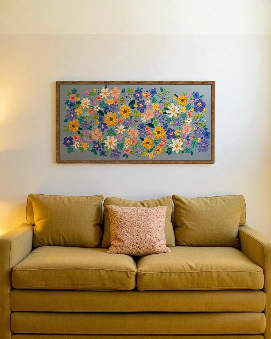 A beige sofa with two cushions, one pink and one beige, beneath a framed artwork featuring paper flowers in multiple colors on a gray background in a living room.