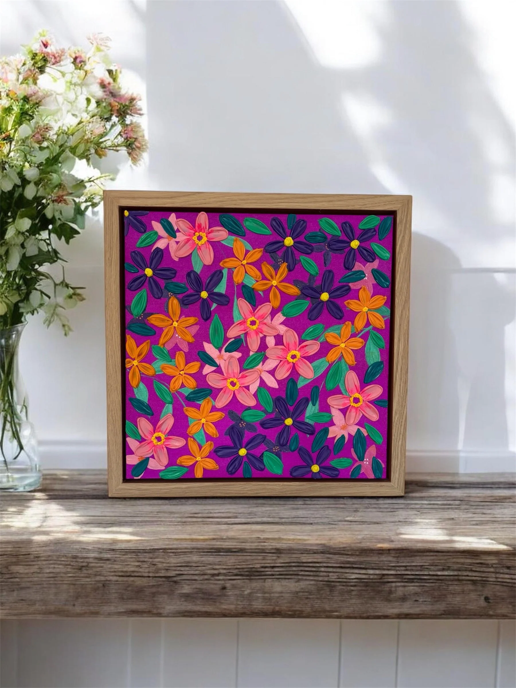 A colorful floral painting featuring pink, orange, purple, and teal flowers with green leaves, framed in natural wood, placed on a wooden surface with a real floral arrangement to the left.