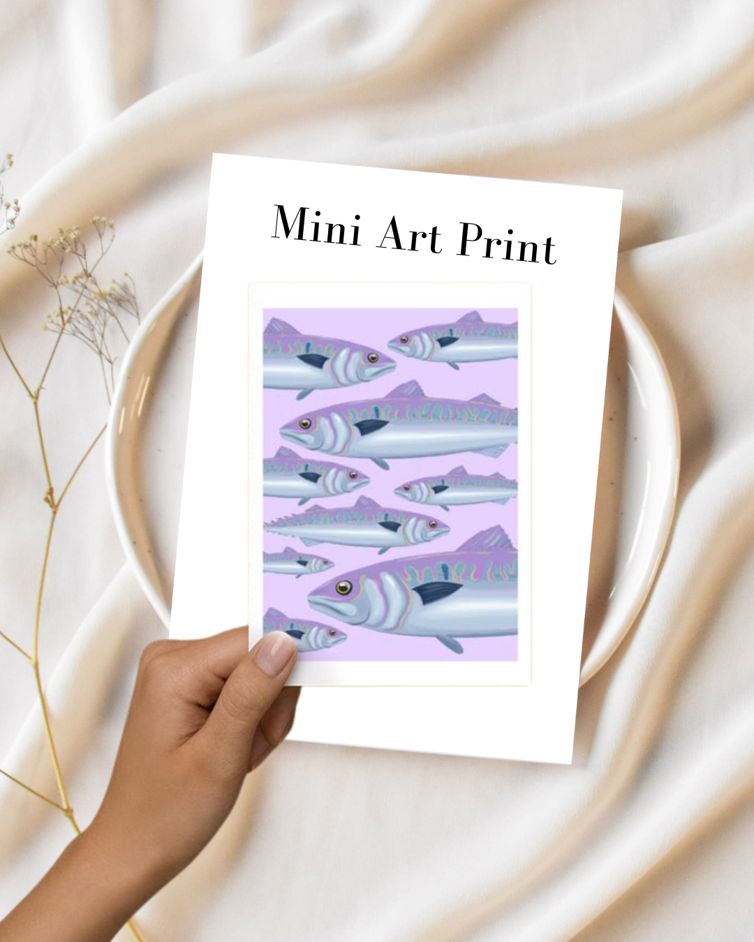 A hand holding a mini art print of illustrated fish with a mountain landscape background, placed over a white ceramic dish and a soft white fabric with dried plant sprigs nearby.