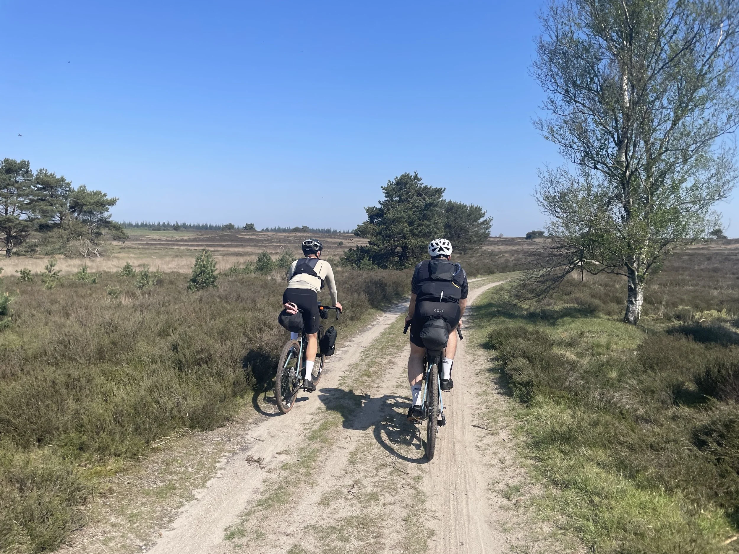 Zwei Radfahrer fahren auf einem unbefestigten Weg durch eine offene Landschaft mit Büschen, Gräsern und einigen Bäumen unter einem klaren blauen Himmel.