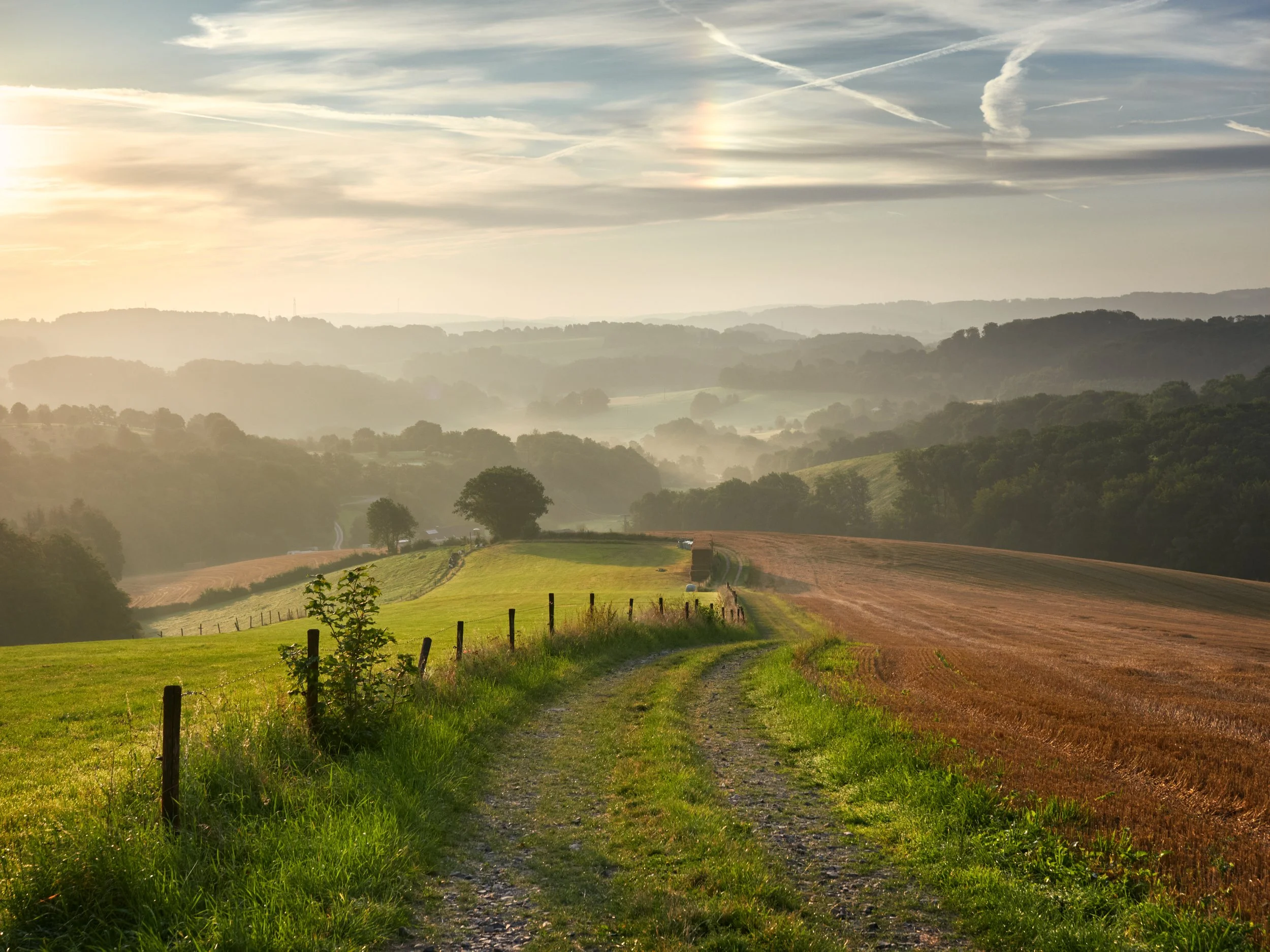 Eine ländliche Landschaft mit einem schmalen Weg, grünem Gras, Feldern und Bäumen, im Hintergrund Hügel und eine bewölkte Himmel mit Sonnenstrahlen und einem kleinen Regenbogen.