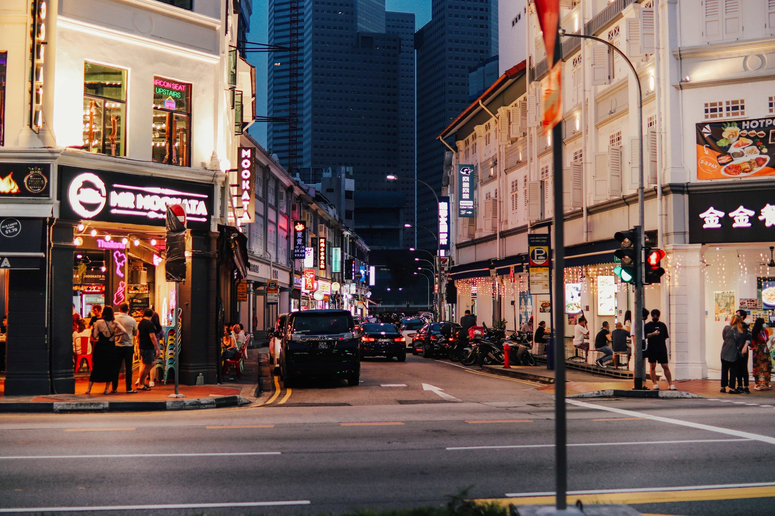 Dark street scene in Singapore with illuminated storefronts and signs, Ignoring the buildings in the background with many high-rise structures. People walking on the sidewalks, parked cars and motorcycles on the street.