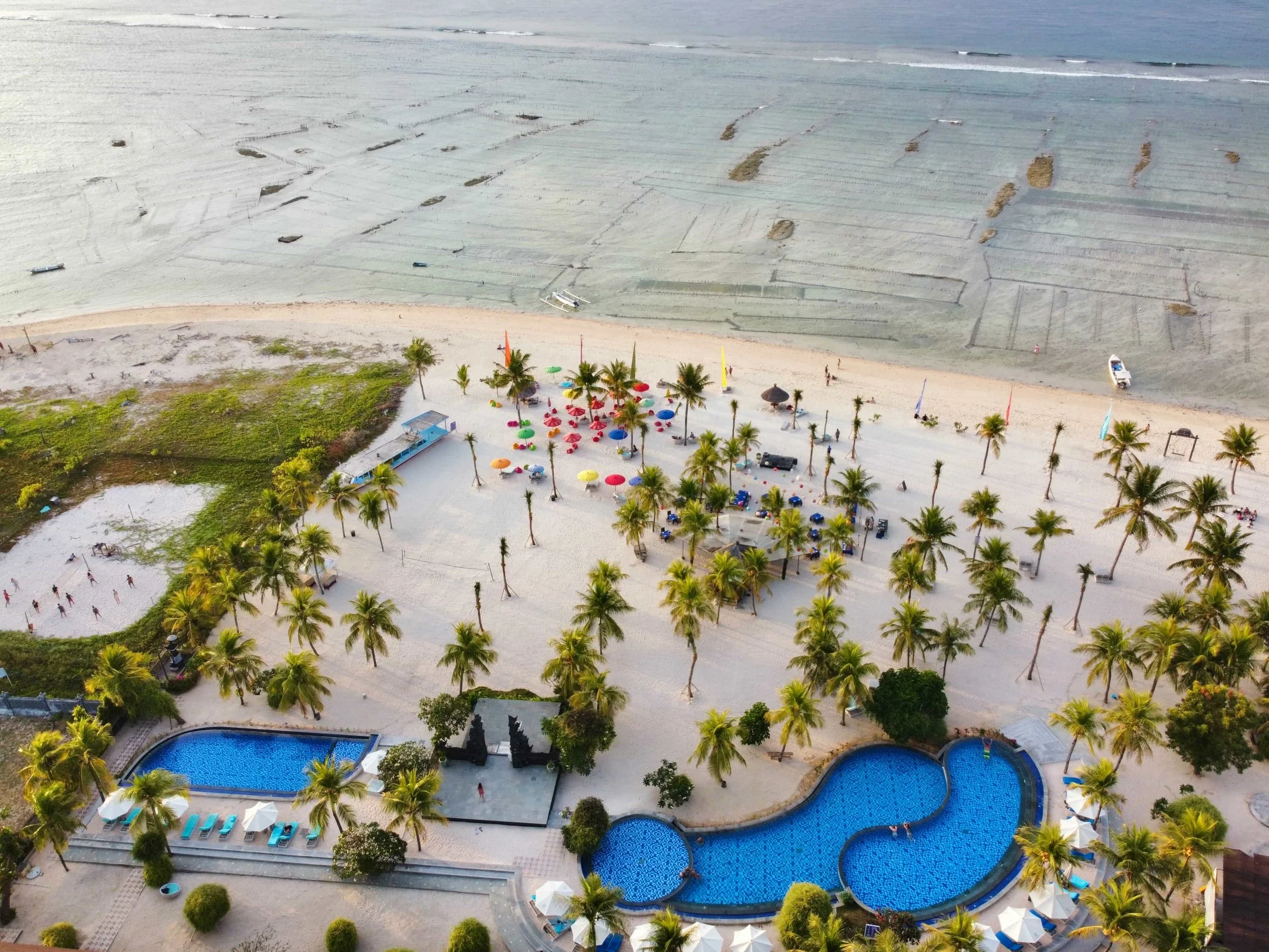 An aerial view of a Filipino beach resort featuring swimming pools, palm trees, and a sandy shoreline with colorful umbrellas, with the ocean in the background.