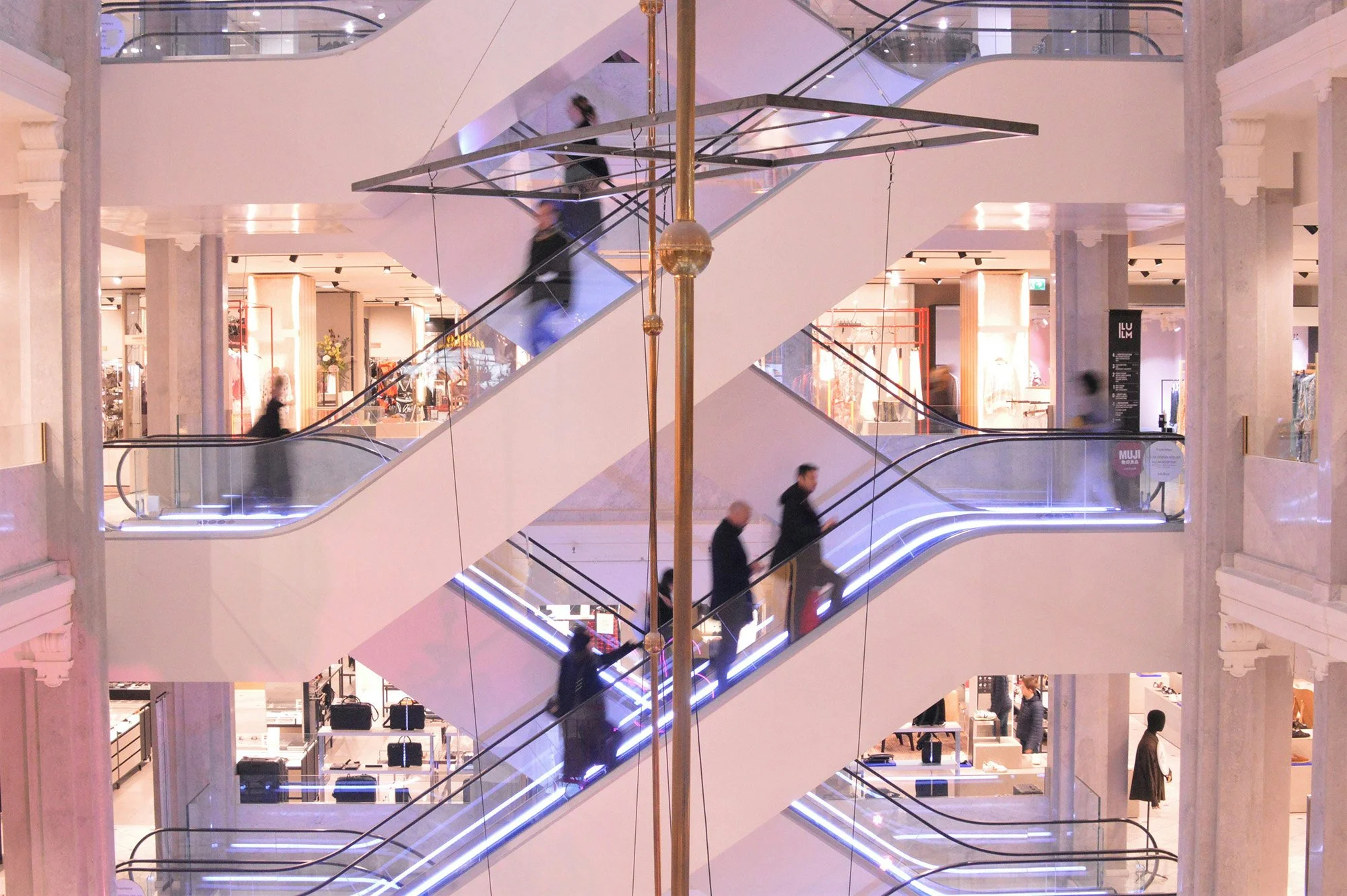 Interior view of a multi-level shopping mall in Asia with a central escalator and wayfinding with people walking on different floors, warm lighting, and retail stores in the background.