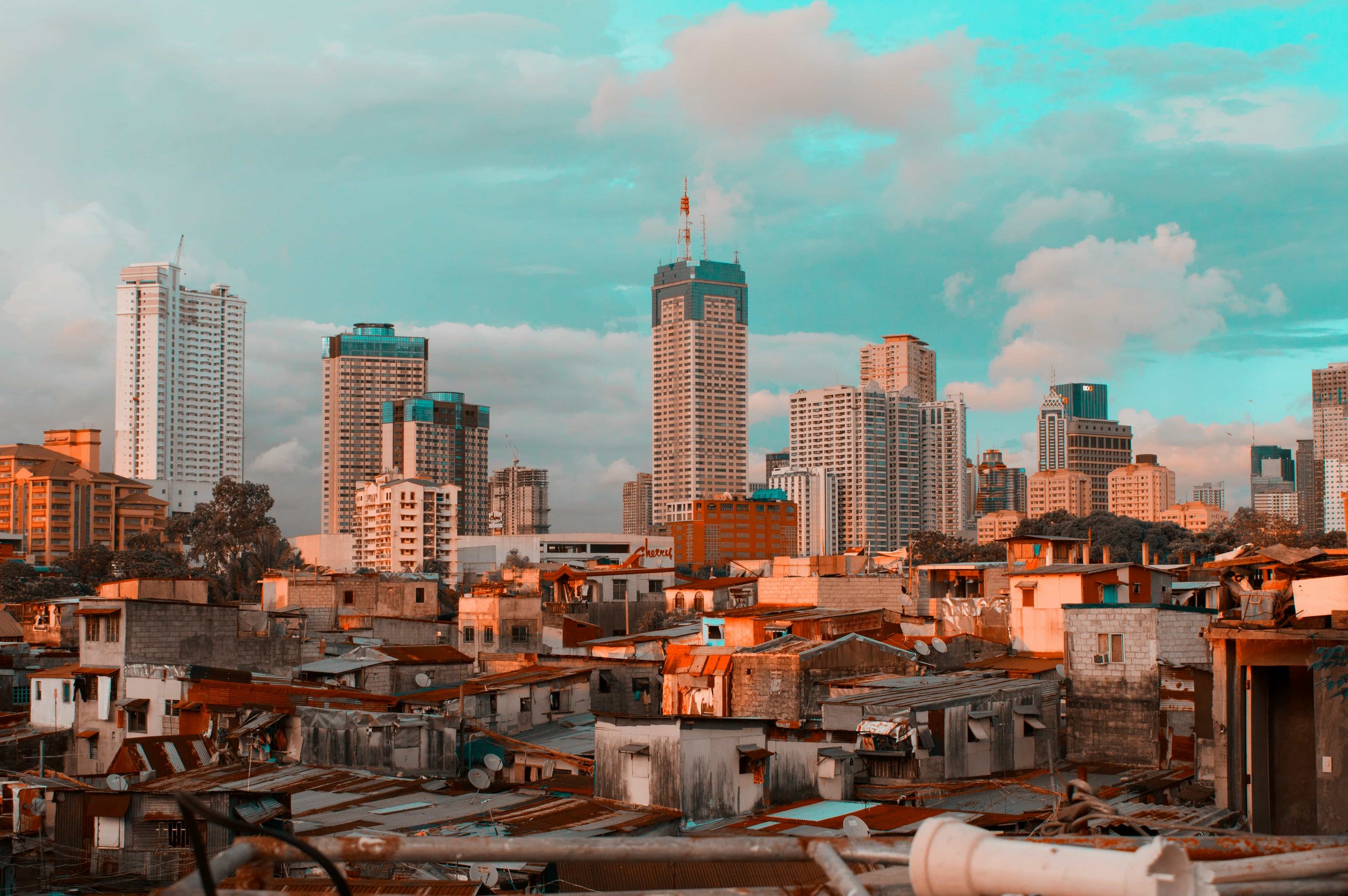 View of the Manila city skyline with tall modern buildings and a foreground of low-rise, makeshift houses with corrugated metal roofs, under a partly cloudy sky.
