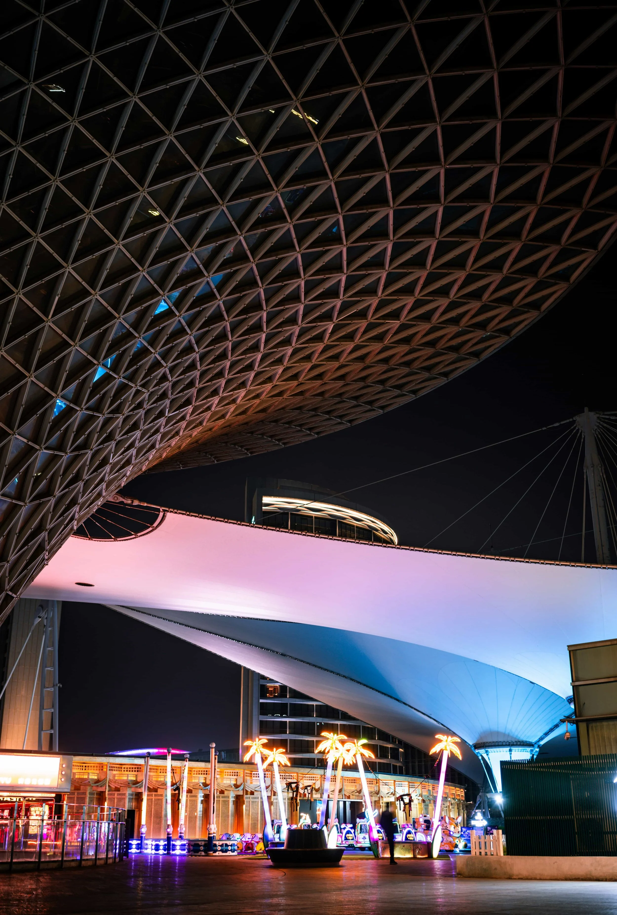 Marina Bay Singapore, Night view of an outdoor amusement park with colorful neon lights, palm tree-shaped lights, and a large curved structure with a grid-like roof.