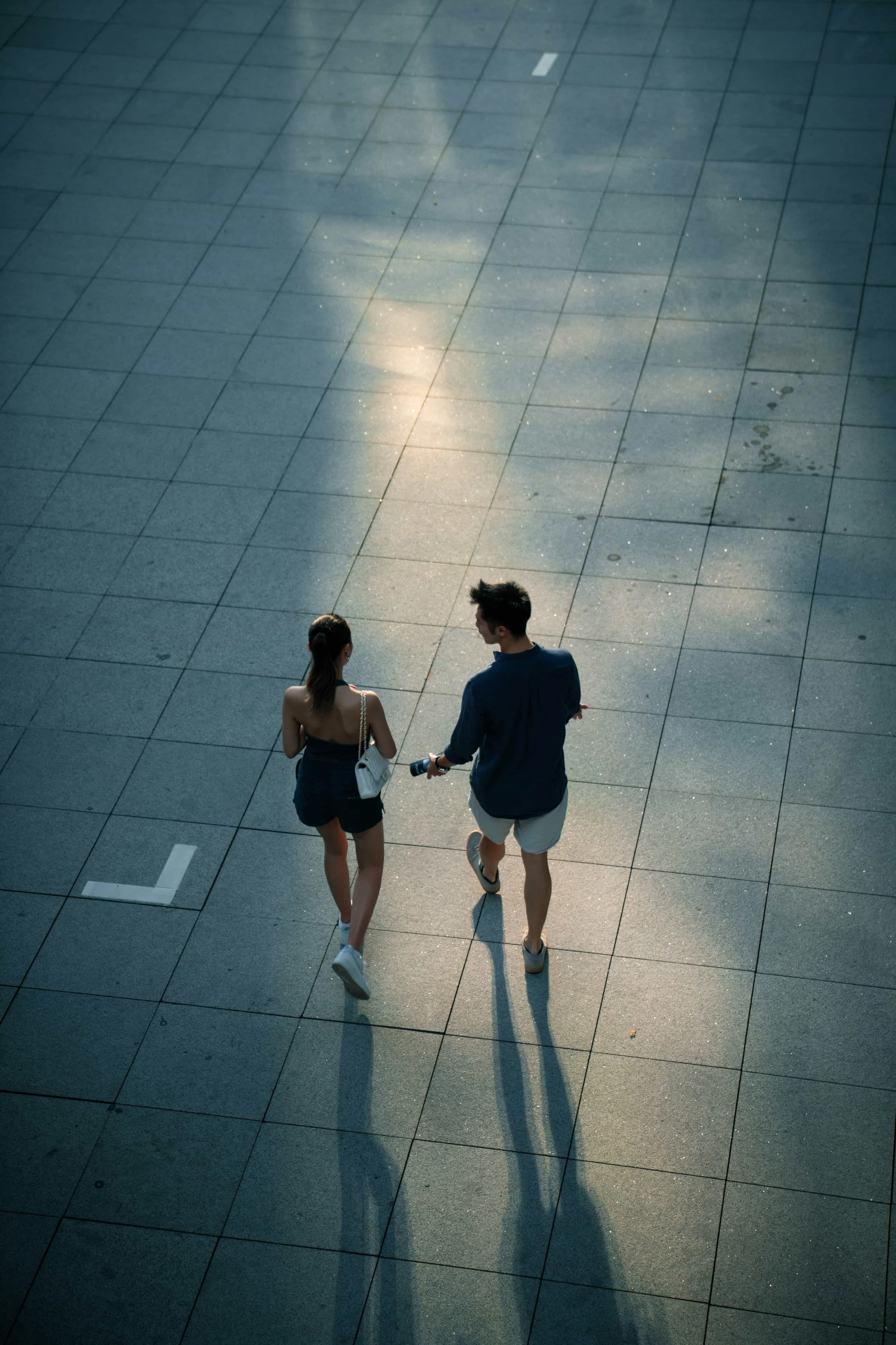 Two people are walking on a tiled surface in Singapore, casting long shadows in the sunlight. One person is a woman with her hair tied back, the other a man exploring their area with wayfinding.