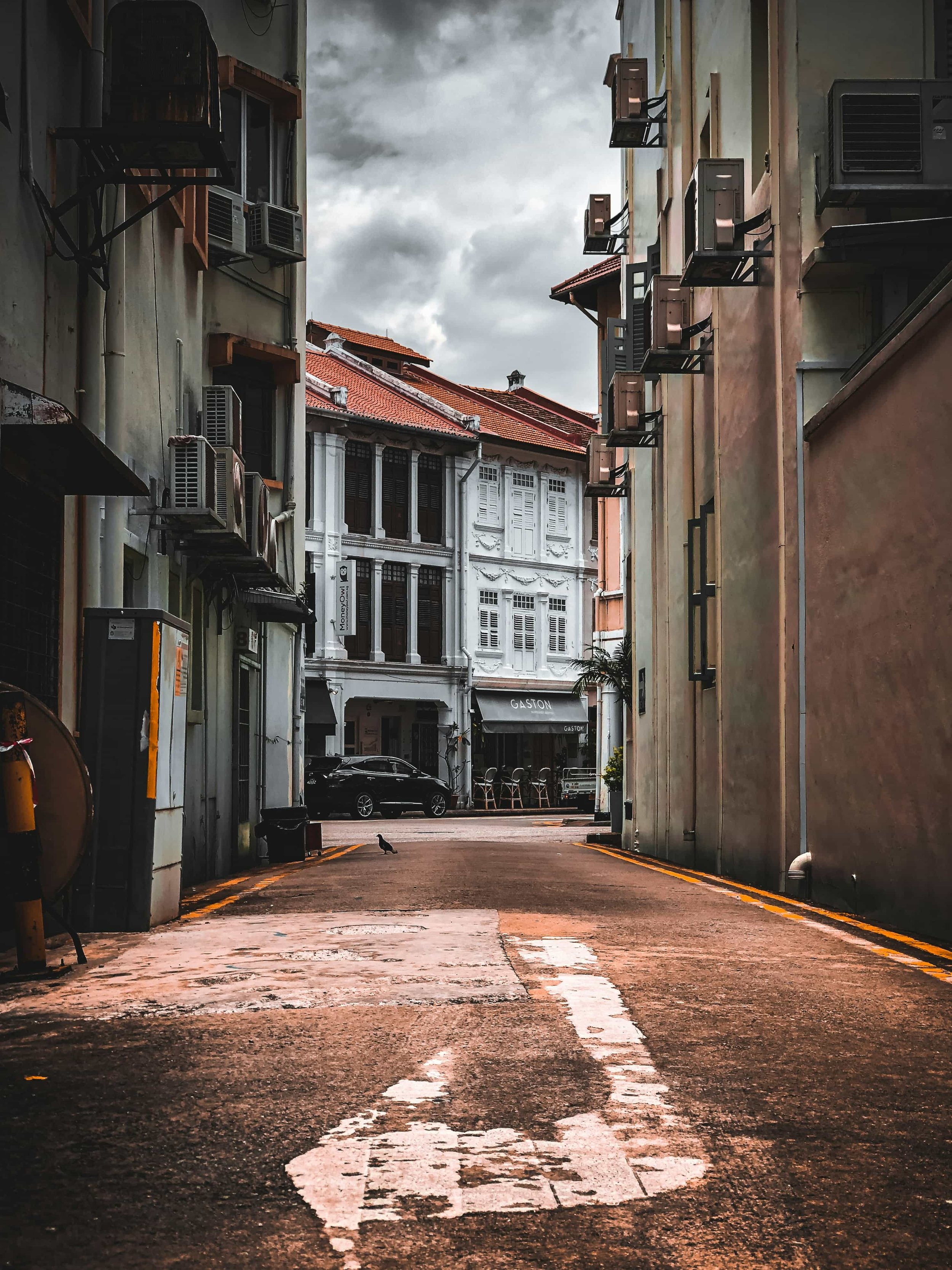 Empty alleyway in Singapore with weathered pavement, flanked by tall buildings with air conditioning units, under a cloudy sky.
