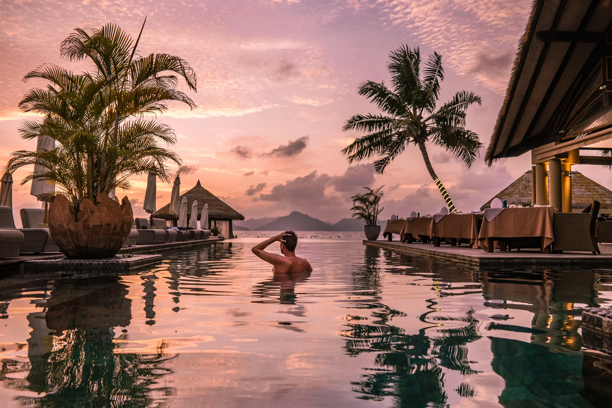 Hospitality. A person swimming in a pool at sunset with palm trees and thatched-roof structures in the background.