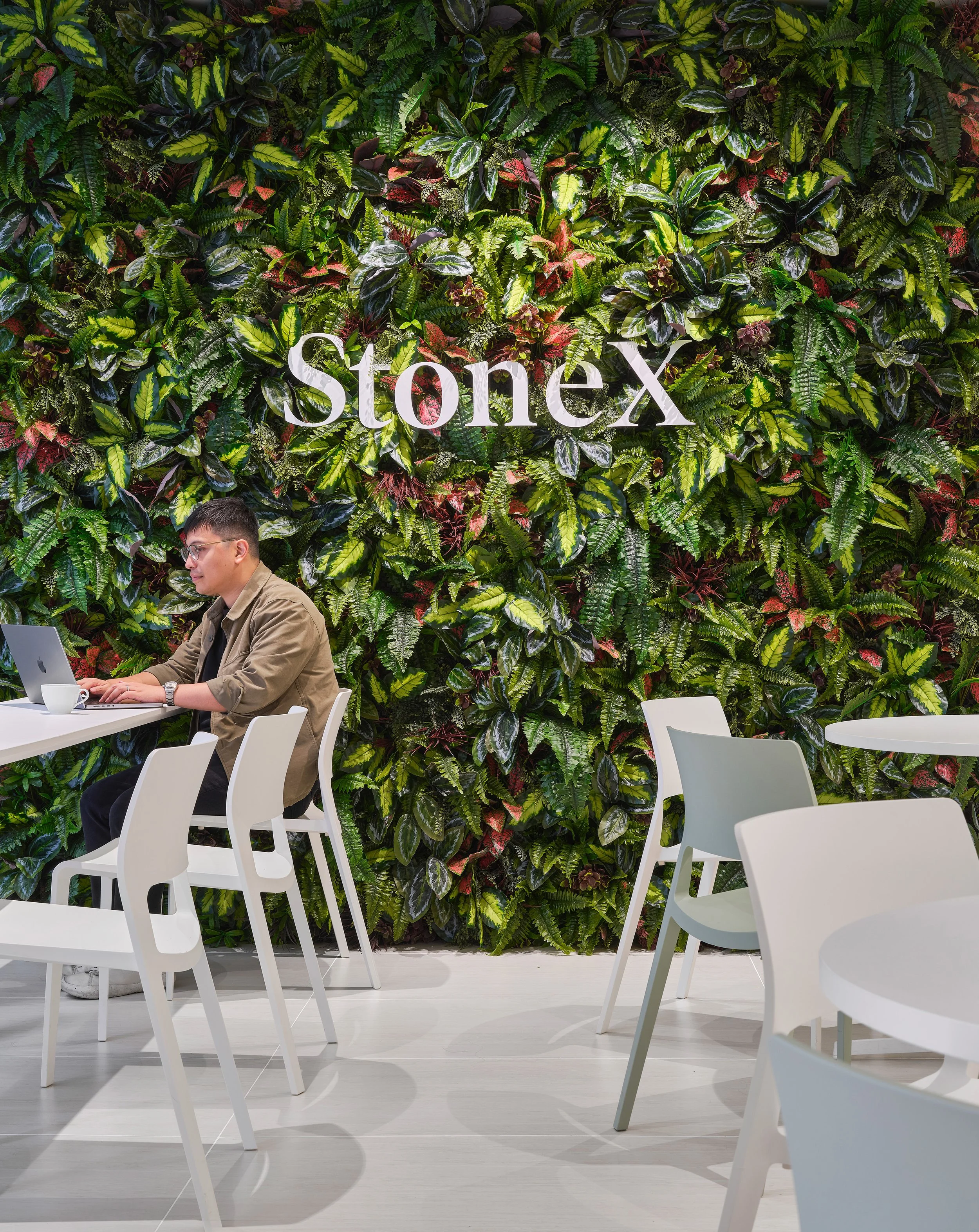 A man sitting at a white table working on a laptop, with a green leafy vertical garden wall behind him displaying the StoneX logo in white letters.