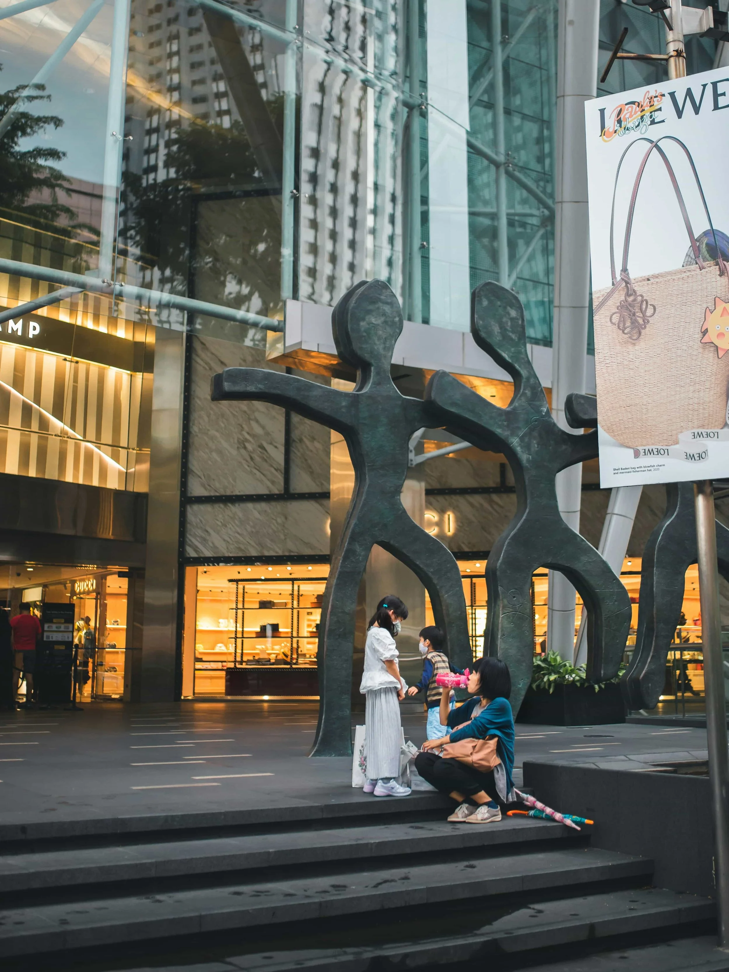 Singapore People, including a woman and children, in a busy urban shopping area near a large metal sculpture and a glass building with stores in the background, some wearing masks.