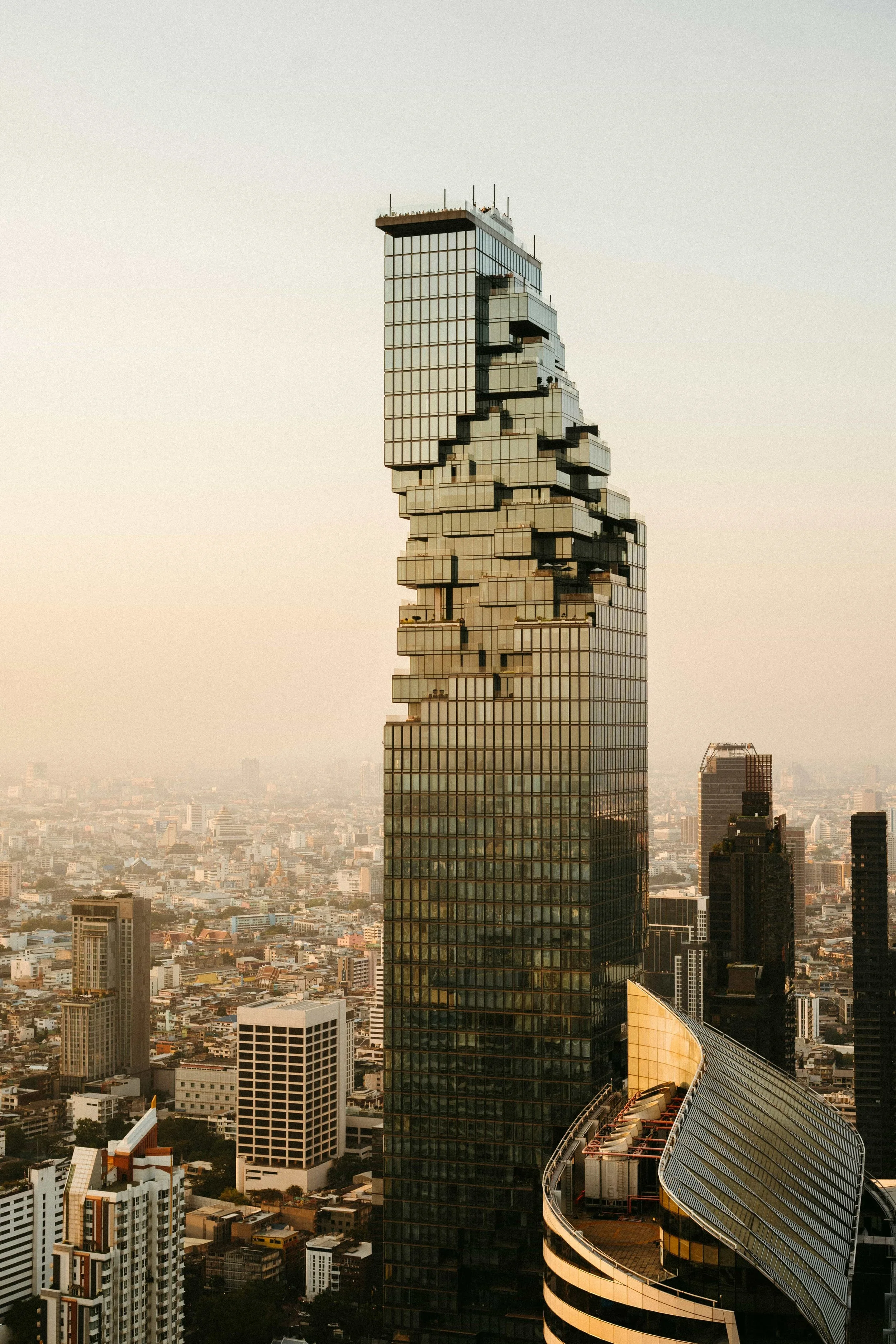 A tall, modern skyscraper in Bangkok, Thailand with a distinctive irregular, stacked design against a cityscape background during sunset.