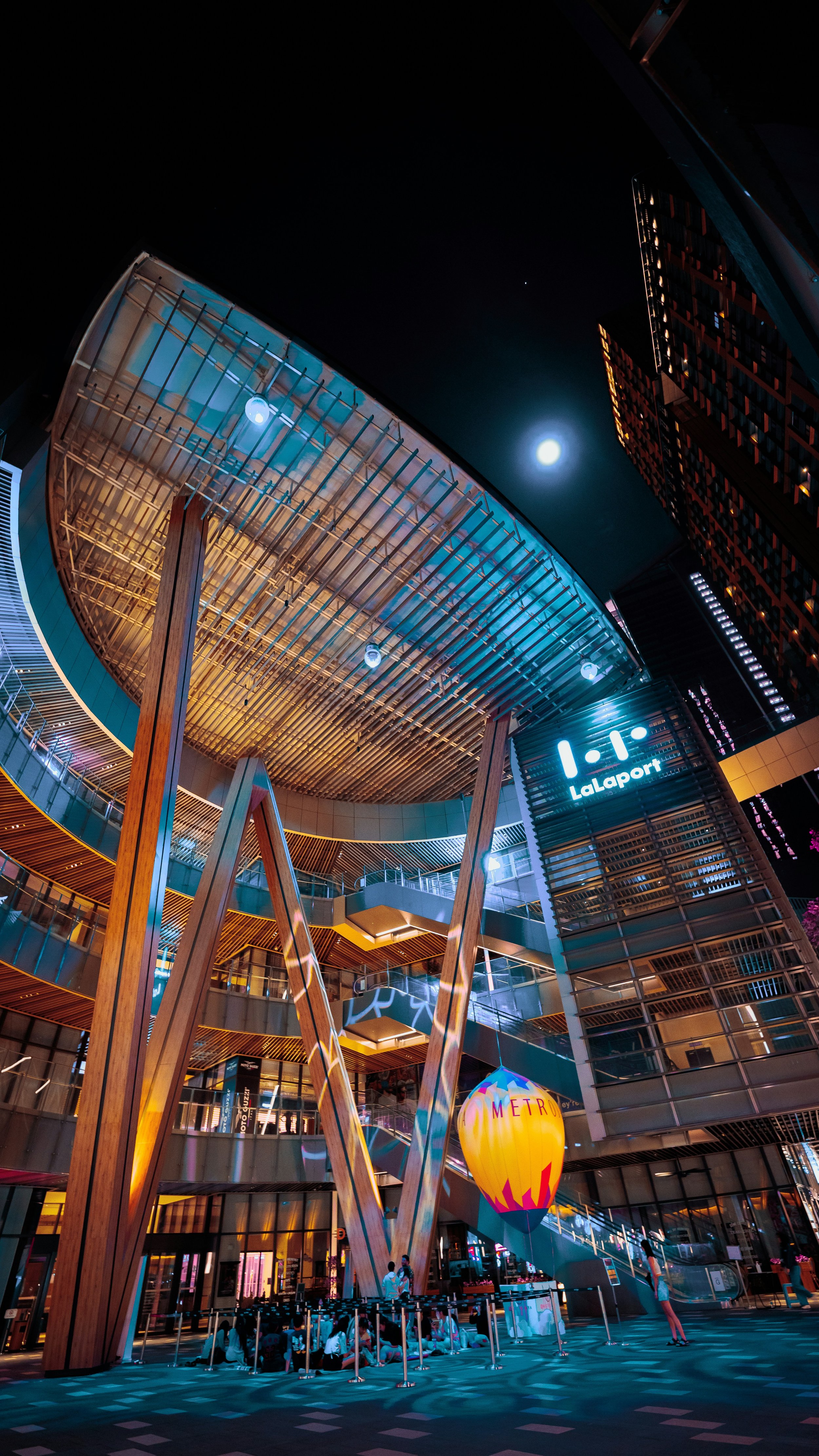 Night view of LaLaport shopping mall entrance with people sitting outside, decorative lighting, a hot air balloon decoration, and tall buildings illuminated in the background.