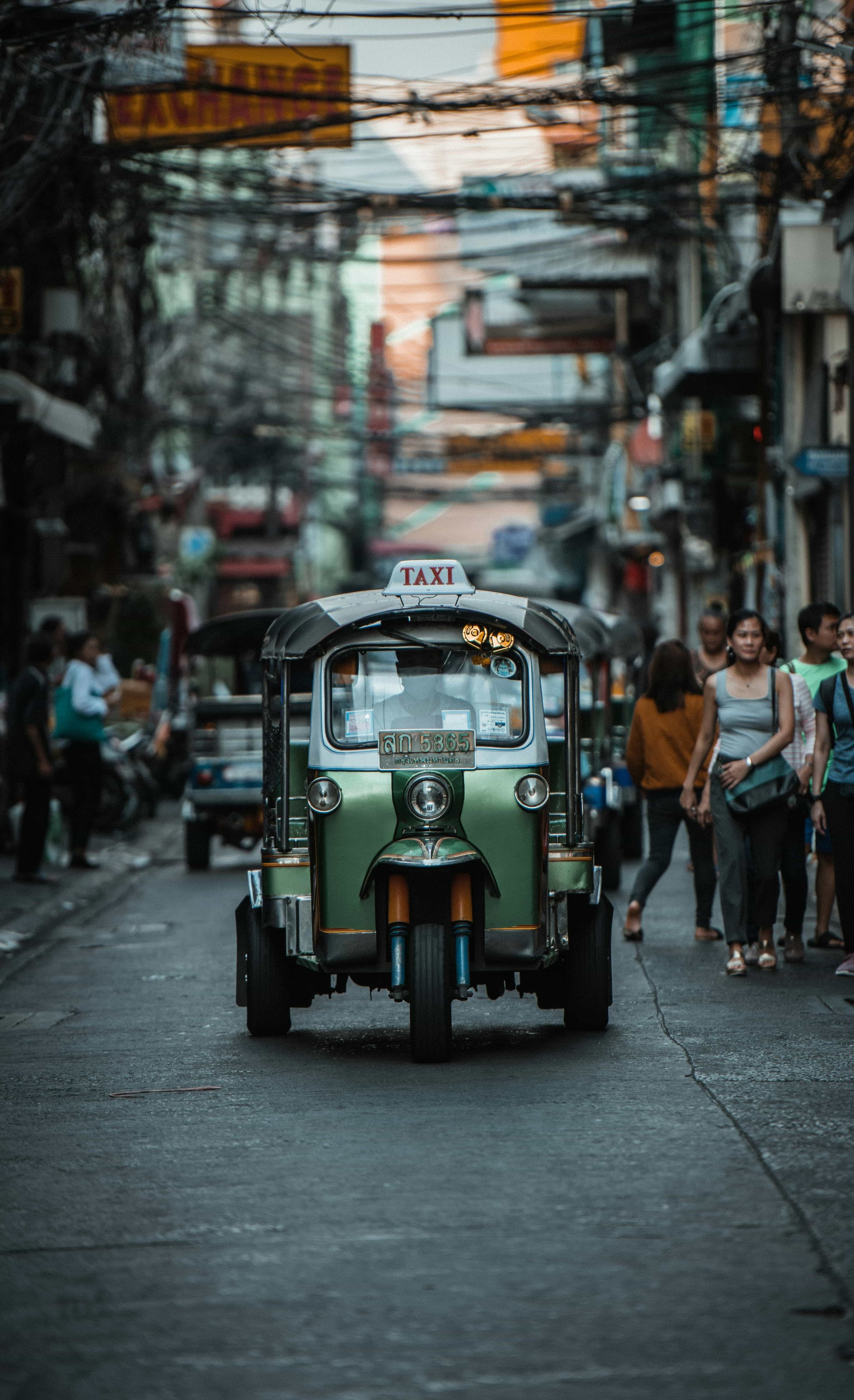 A green tuk-tuk, a three-wheeled motorized vehicle, driving down a busy Thailand street with pedestrians walking on the sidewalk. Buildings and power lines are visible in the background.