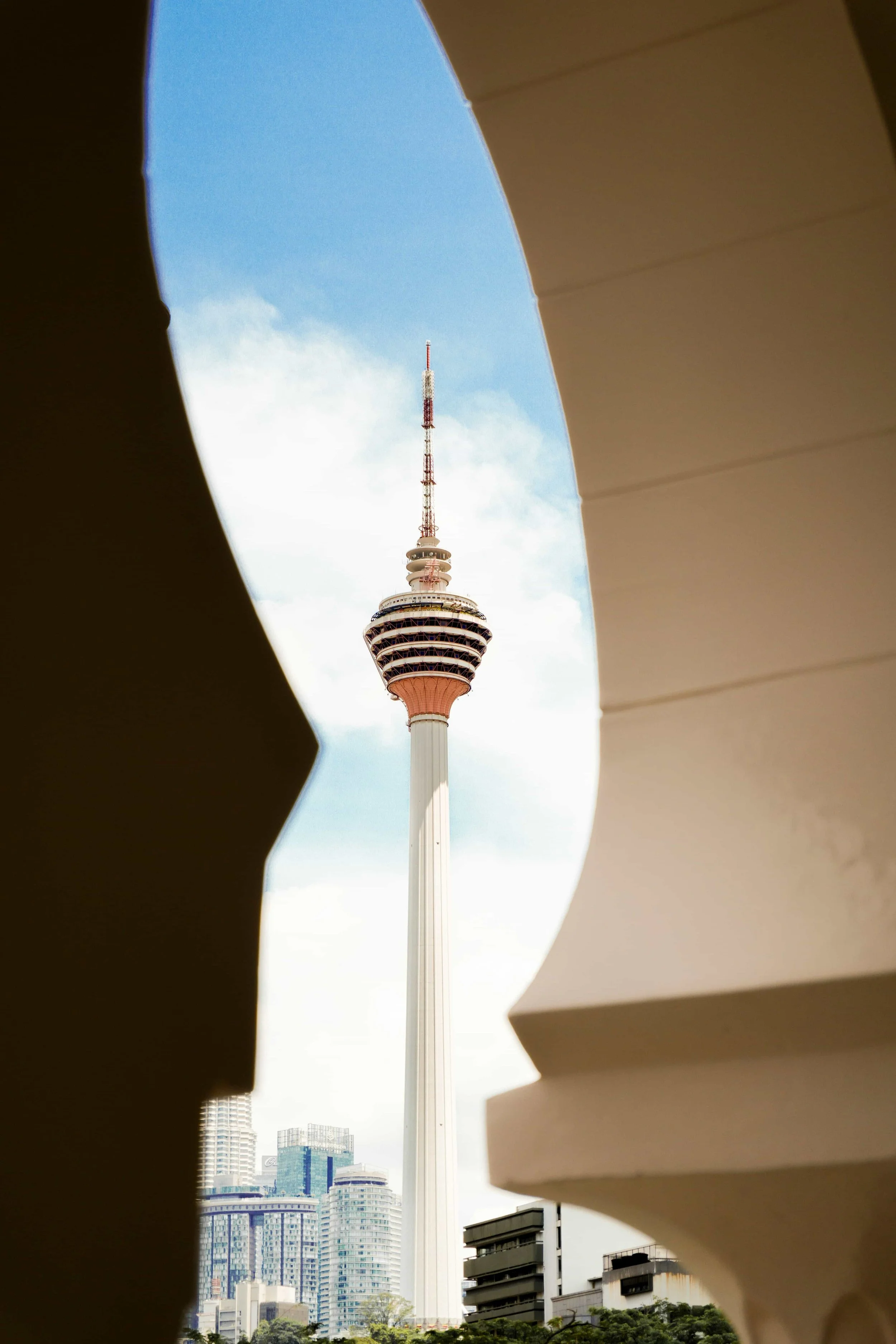 A view of a tall telecommunications tower in KL through an opening in a beige building or structure with curved edges, with a cityscape of modern high-rise buildings in the background and a blue sky with some clouds.