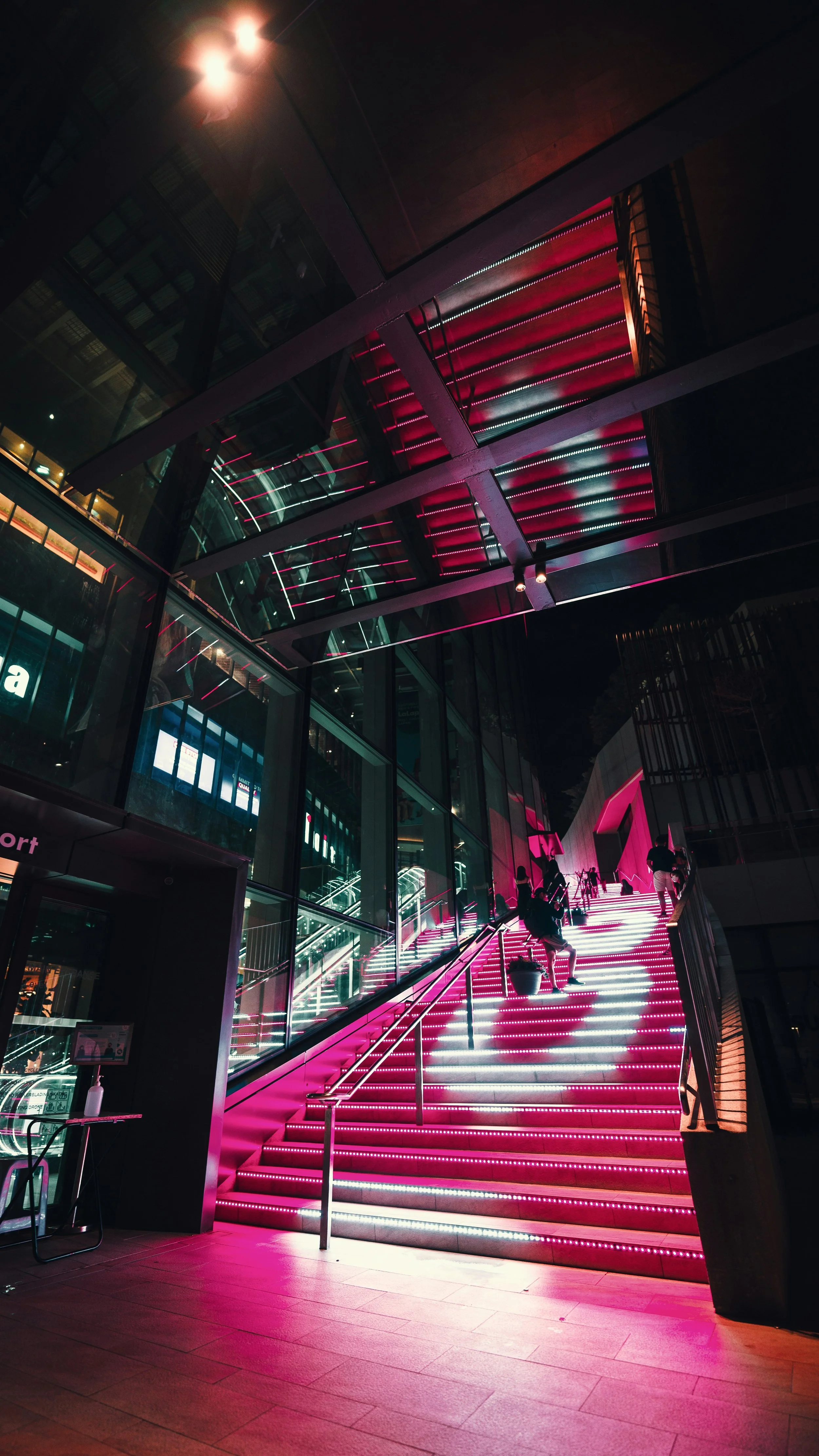 Night view of a modern building entrance with illuminated pink stairs and glass walls, people ascending stairs, and reflective ceiling with pink and white lights.