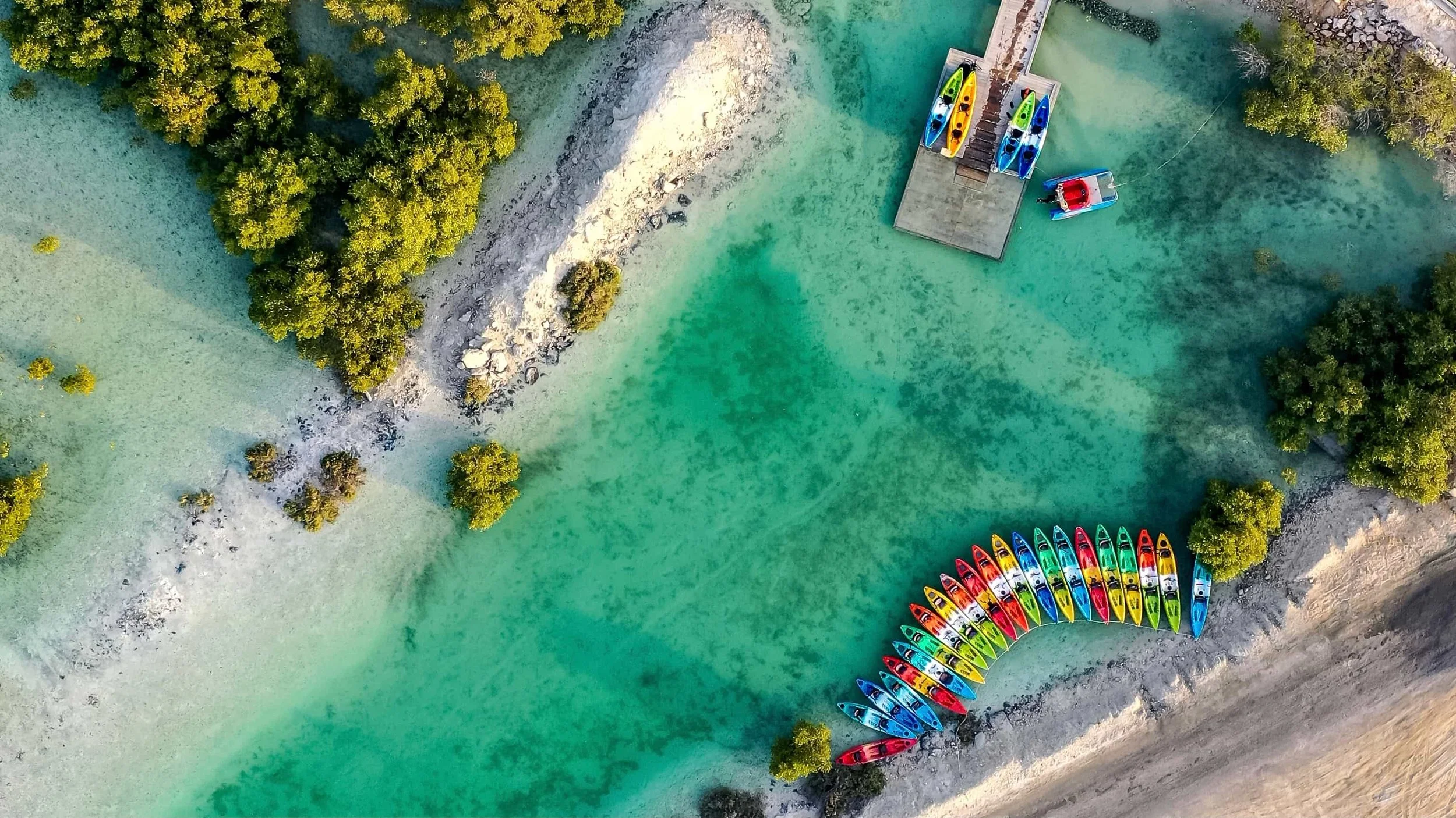 An aerial view of a small dock with colorful kayaks in the Philipines lined up along the shore and in the water, surrounded by clear turquoise water, trees, and rocks.