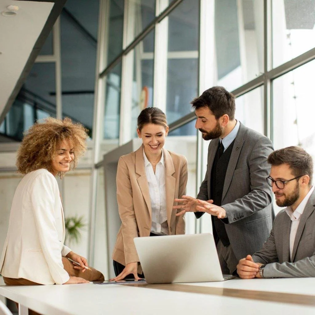 A group of five diverse professionals gathered around a laptop in a bright office, engaged in a discussion and smiling.