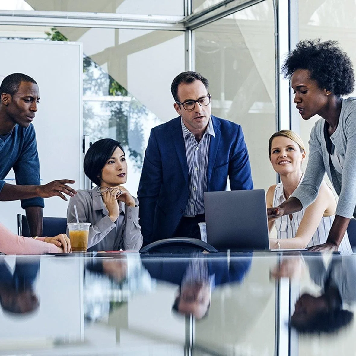 A diverse group of five professionals gathered around a conference table in a modern office, engaged in a discussion while looking at a laptop and a woman points at the screen.