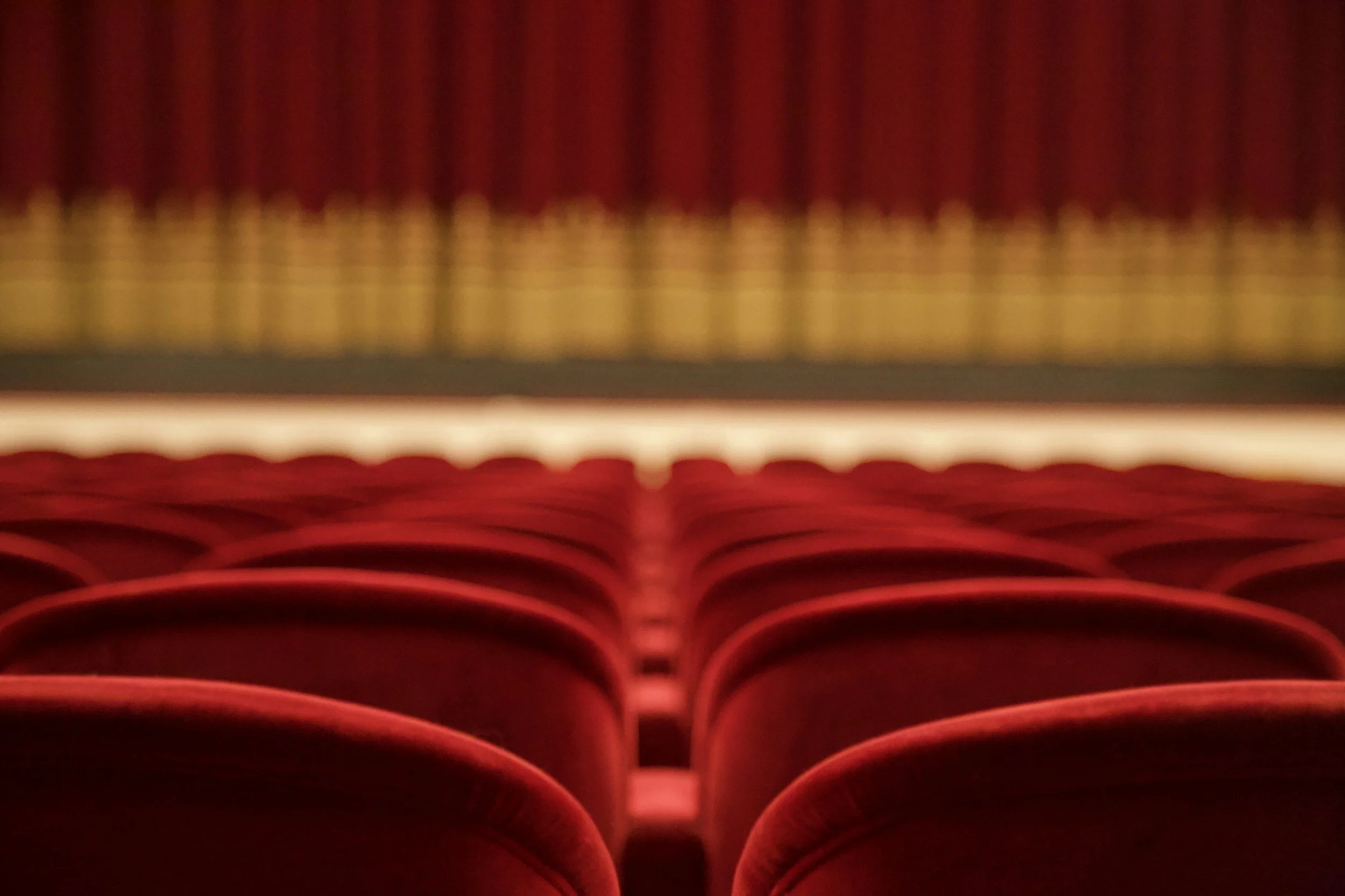 Immagine di un teatro vuoto con sedie rosse, vista dal palco con tende rosse sullo sfondo.